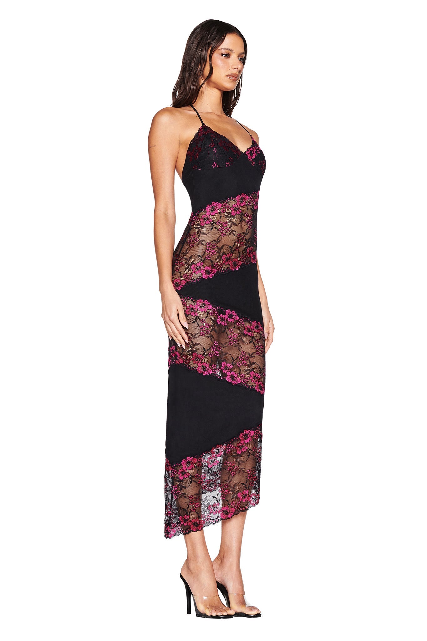 A woman wears the I.AM.GIA PALOMA MIDI DRESS - BLACK, a sleeveless body-con halter style with sheer diagonal lace panels, pink floral details, and an asymmetrical lace hem, paired with black high heels against a white background.