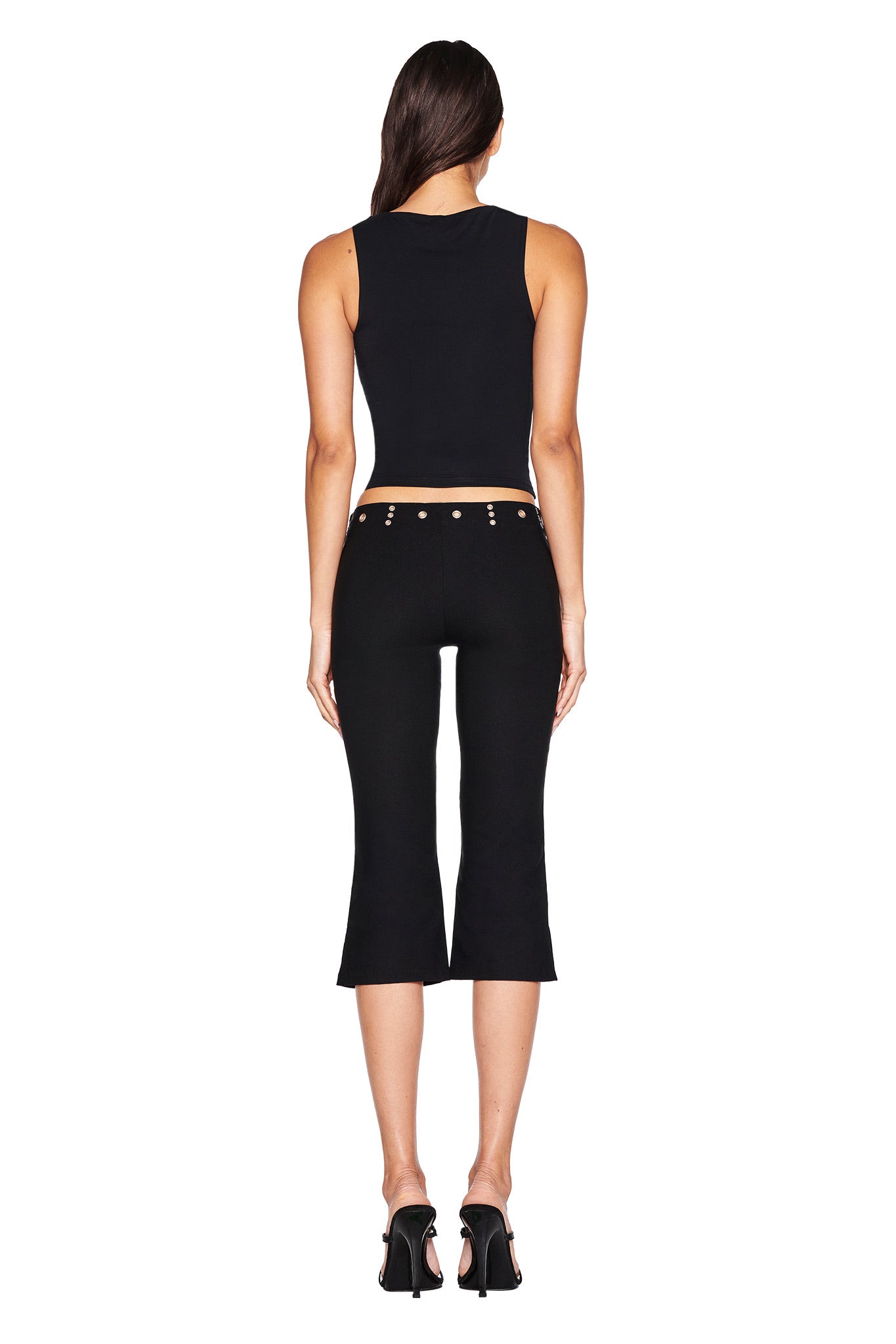A woman faces away, wearing I.AM.GIA’s SHARAH CAPRI - BLACK, a slim, low-rise black capri with button details and mid-stretch fabric, styled with a sleeveless crop top and black heels against a plain white background.