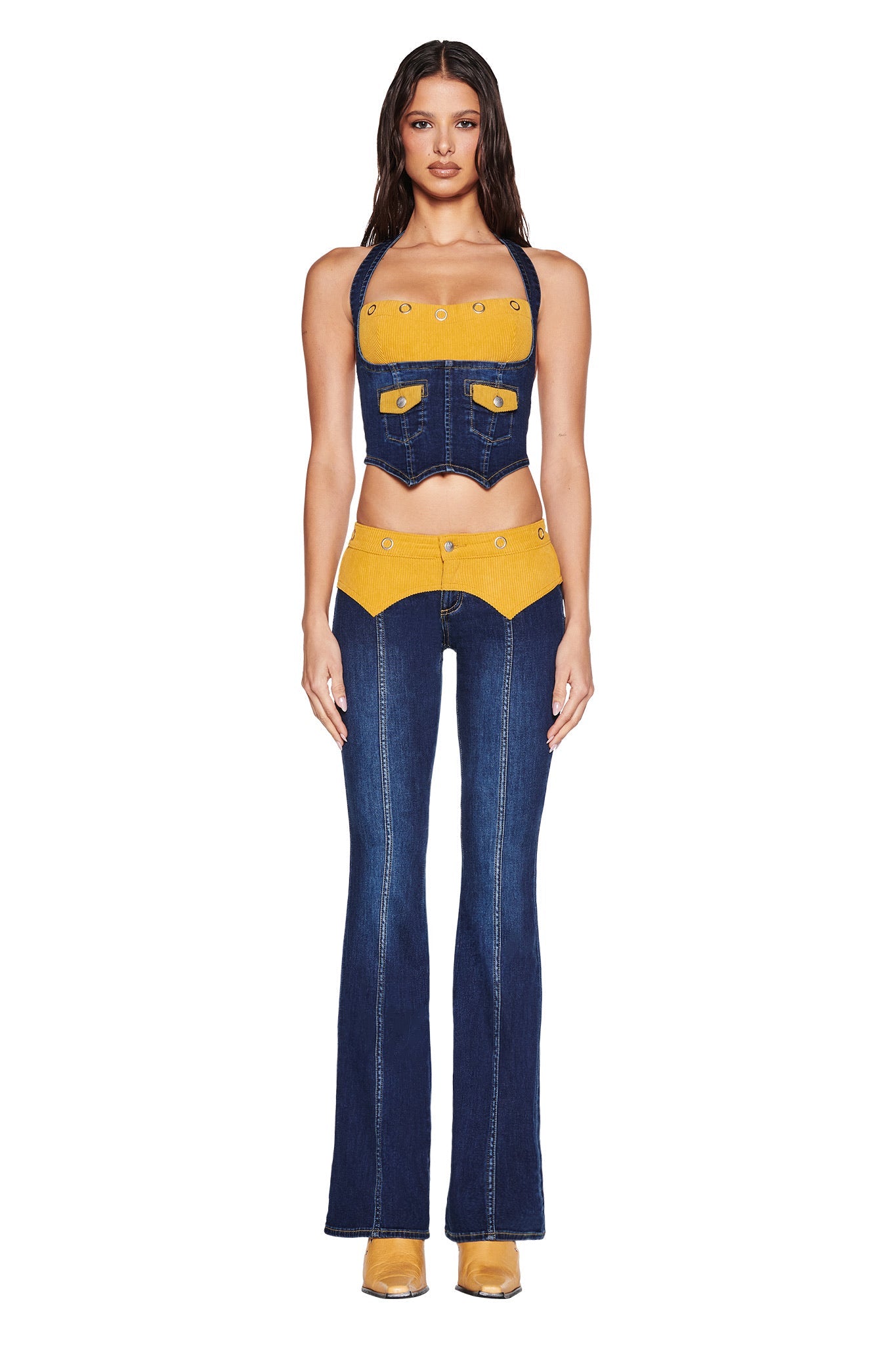 A woman faces forward in the I.AM.GIA TOMMIE JEAN - DARK WASH set, featuring a unique denim and yellow two-piece with corduroy waistband and flared pants. She wears tan shoes and her wavy hair flows against a white backdrop.