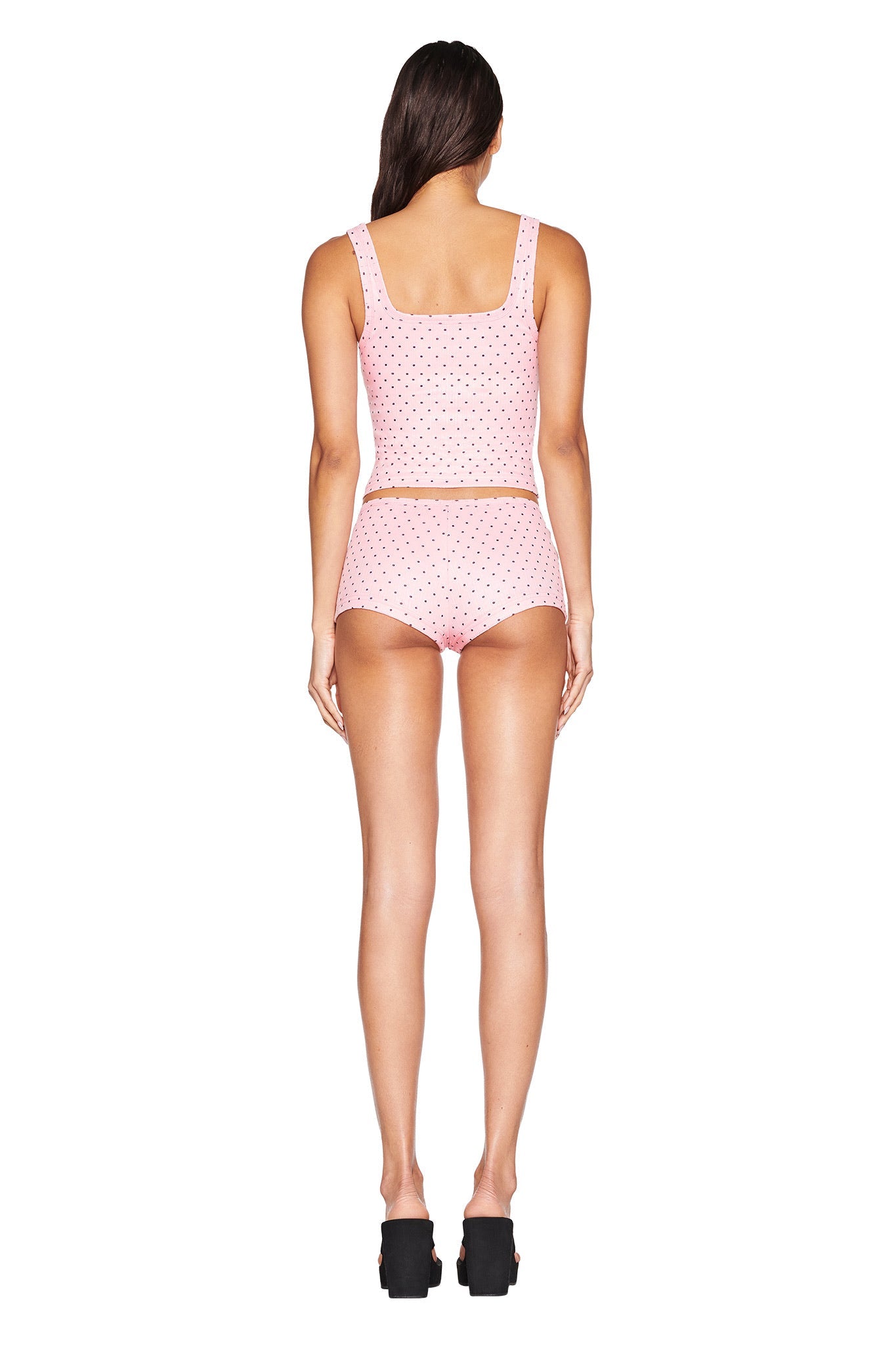 A woman with long brown hair faces away, wearing the I.AM.GIA LAUREL SHORT in light pink, a matching crop top, and black platform shoes against a white background.