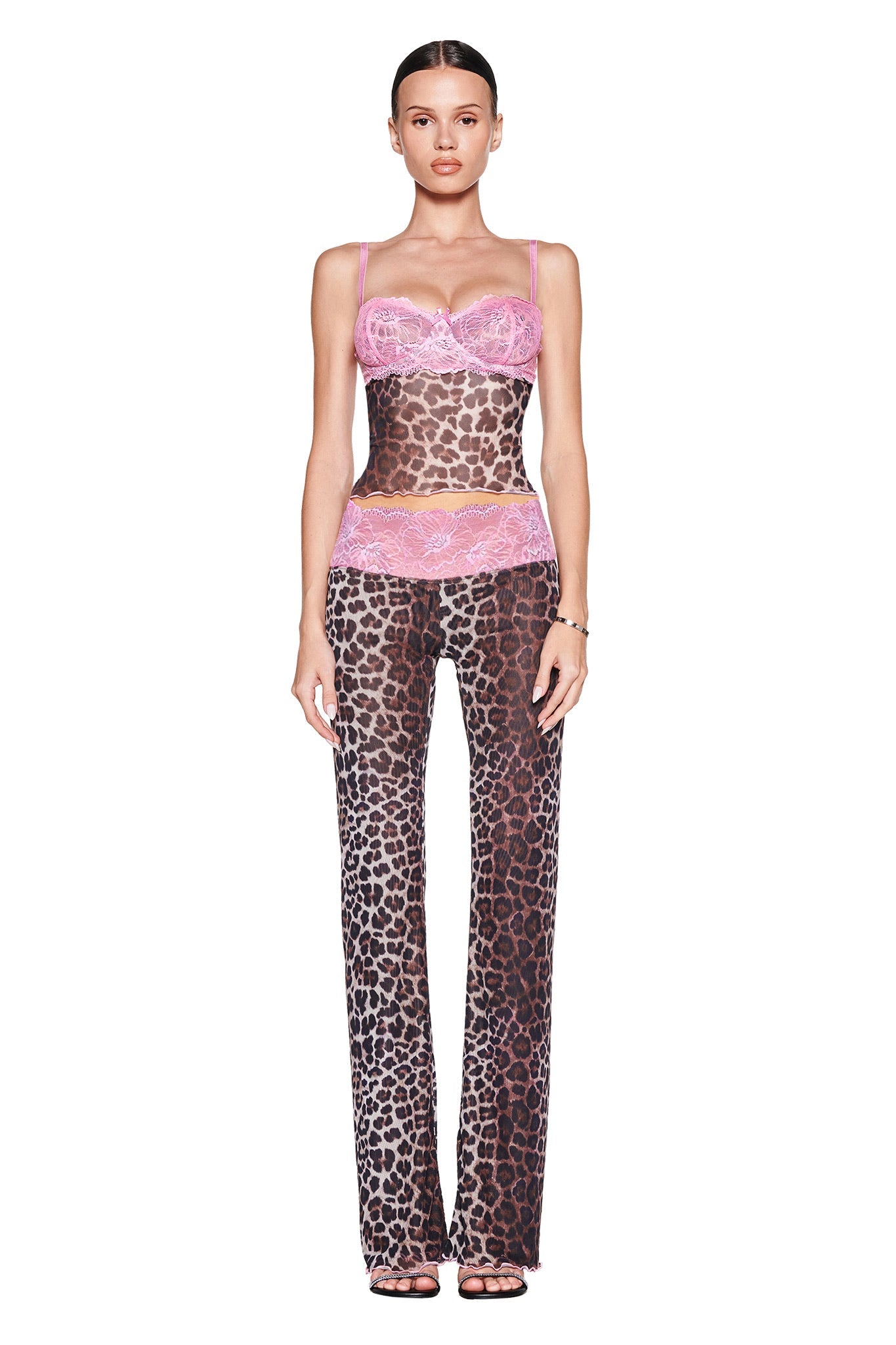 A woman faces forward in I.AM.GIA's SILO PANT - CLASSIC LEOPARD and a sheer top with pink lace accents. Her hair is pulled back, and she wears open-toed heels against a crisp white background.