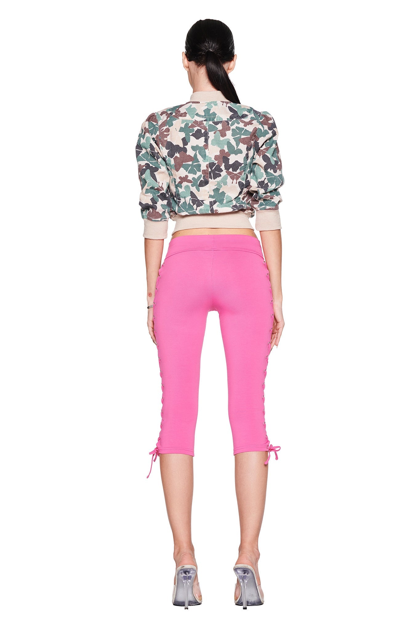 A person with long dark hair tied back wears I.AM.GIA's TILA CAPRI in hot pink, featuring lace-up detailing on the sides, paired with a cropped camouflage jacket and silver high heels against a plain white background.