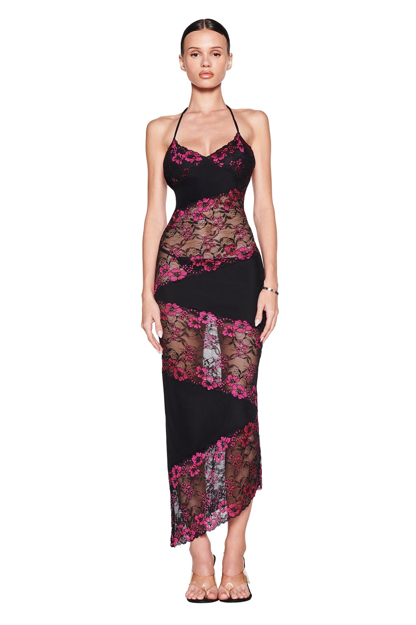 A woman faces forward in the I.AM.GIA PALOMA MIDI DRESS - BLACK, a body-con halter style featuring sheer black lace, pink floral accents, and an asymmetrical hem. She has slicked-back hair and wears sandals.