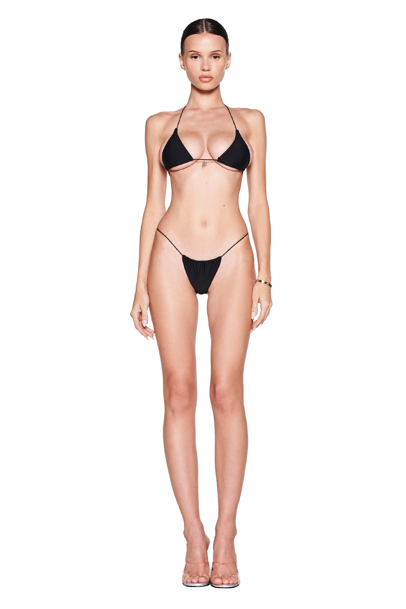 Against a white background, a person showcases the I.AM.GIA black halter neck Bambi Bikini Top, featuring silver 'GIA' hardware and string sides. Their hair is sleekly pulled back, and they pair it with transparent high heels for an elegant touch.