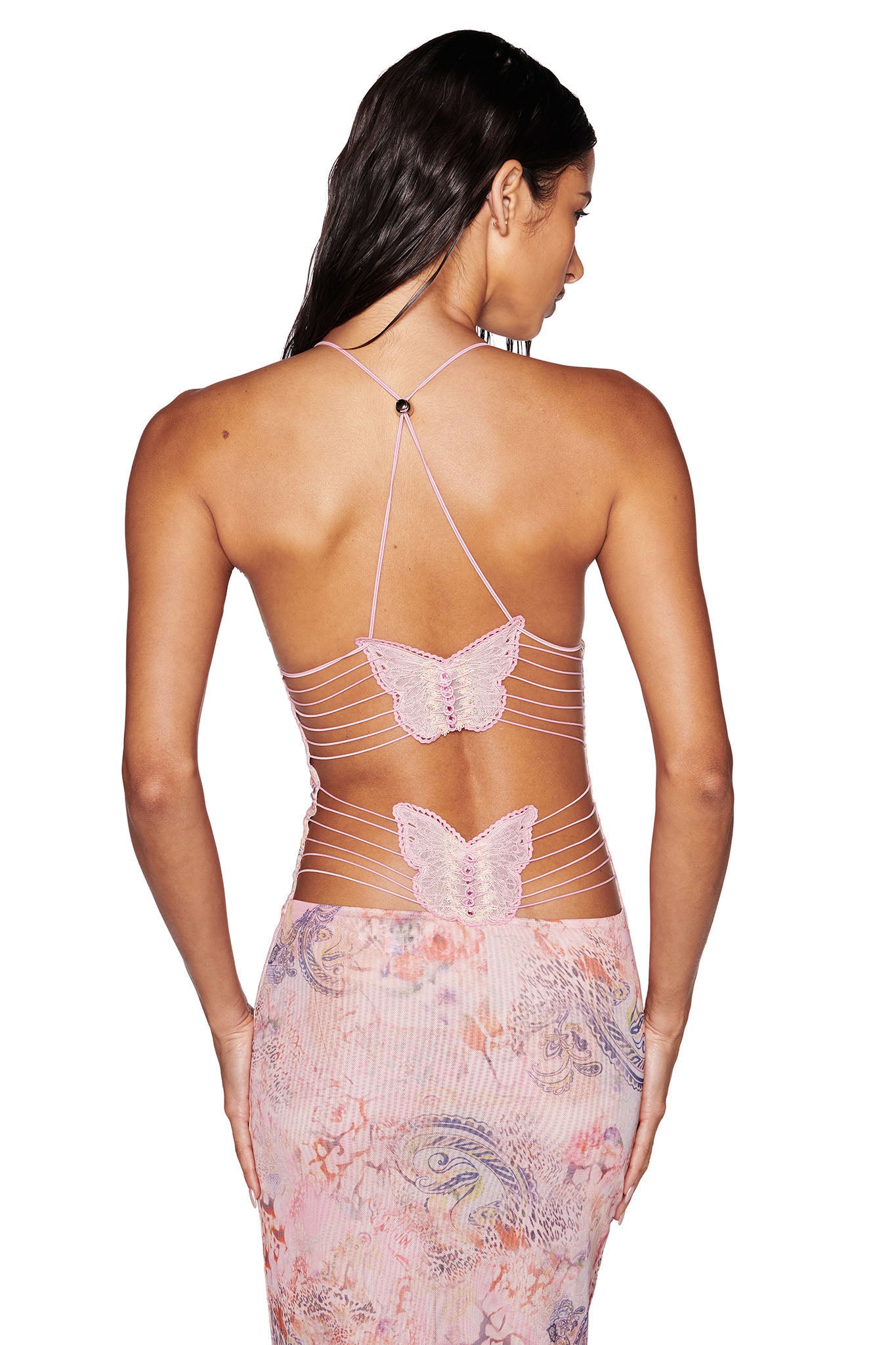 A woman is shown from behind, wearing the I.AM.GIA ELLERY DRESS in pink with a flora paisley pattern. The dress features crisscross straps with two butterfly embellishments and an exposed open back look. She has long dark hair cascading down her back.