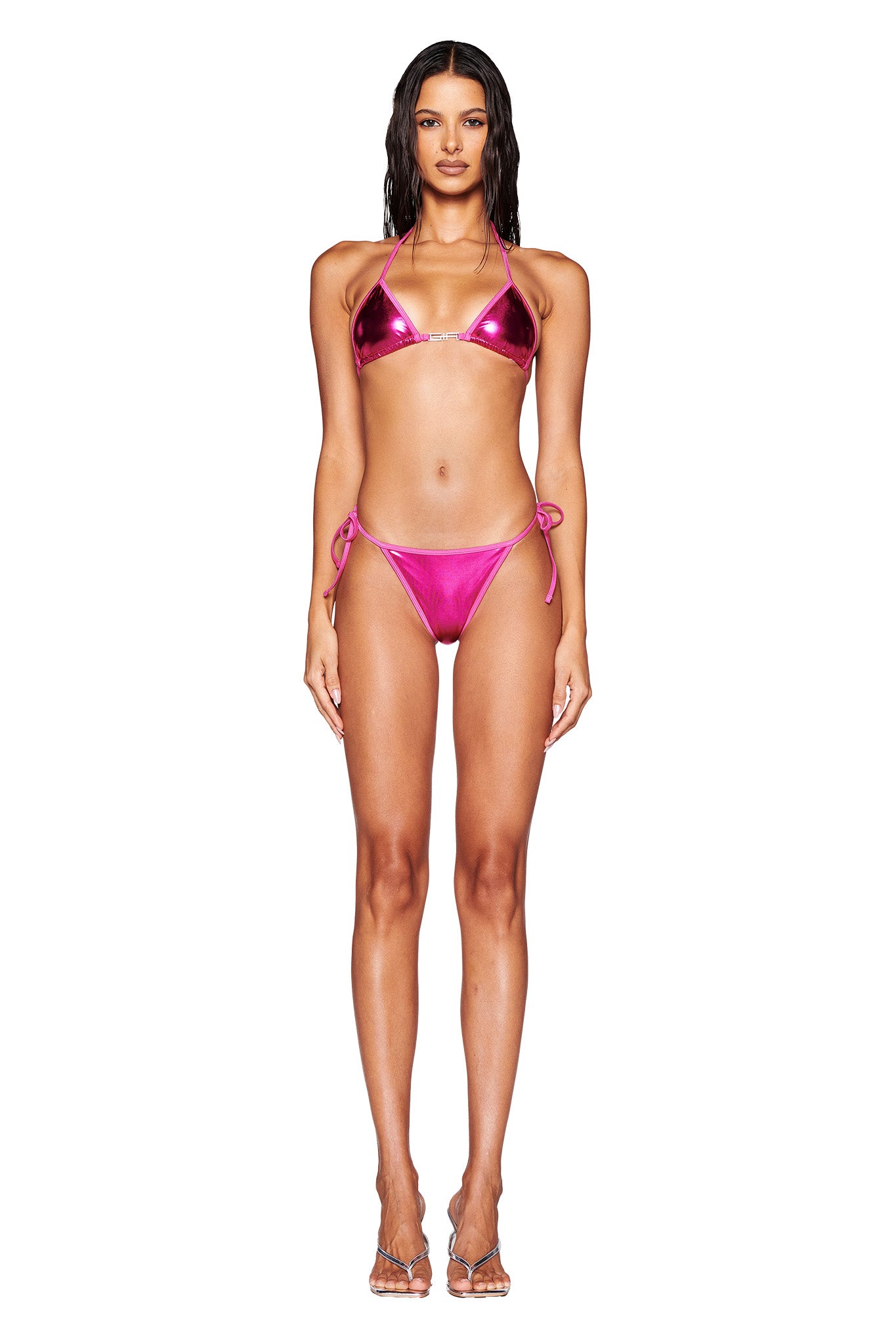 A woman stands against a plain white background wearing the shiny Hot Pink ALOHI BIKINI BOTTOM from I.AM.GIA. Her low-rise bikini bottoms feature cheeky strings on the sides, and the top has thin straps that tie around her neck and back. She also wears clear high-heeled sandals, with her hair wet and slicked back.