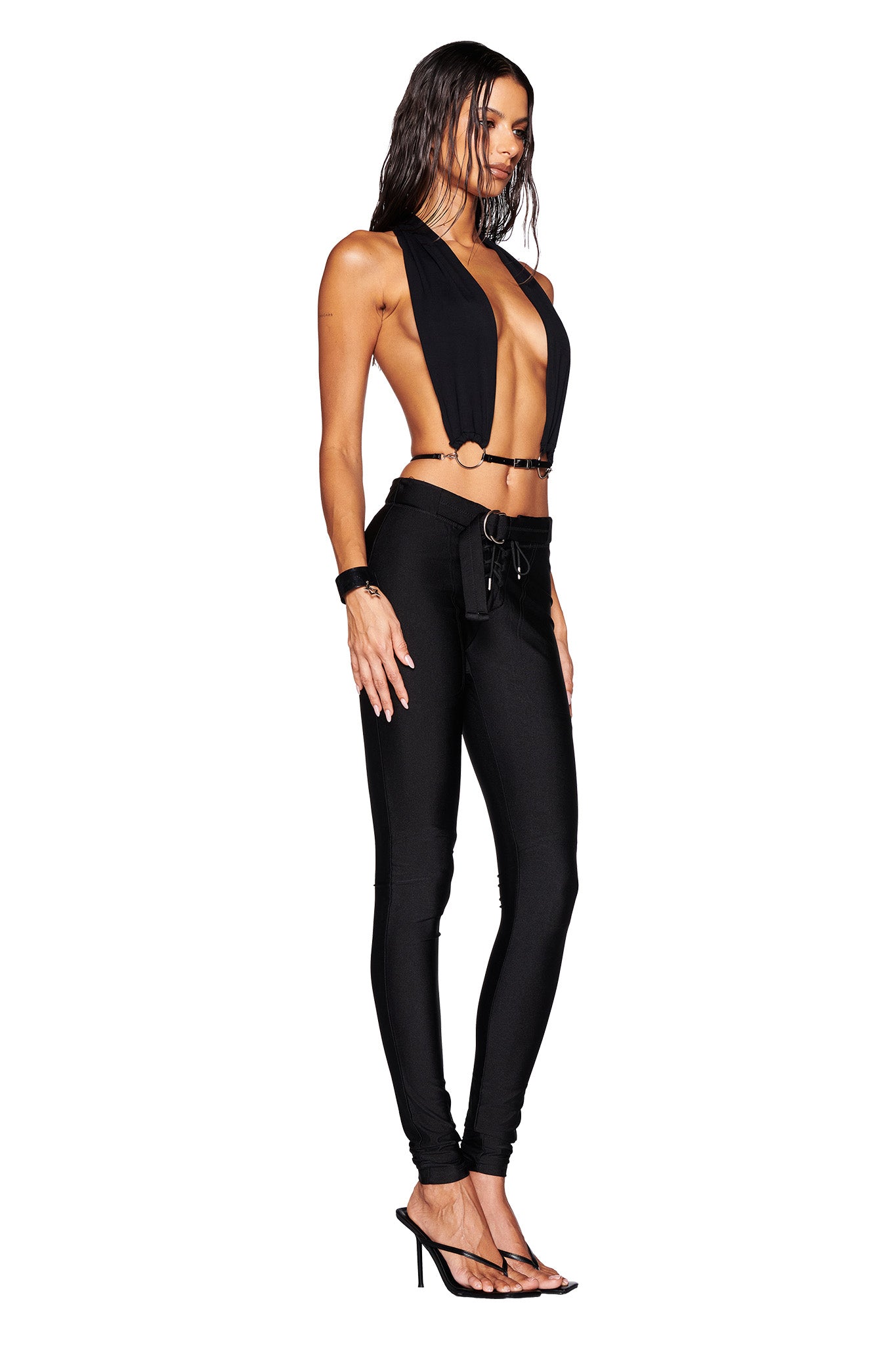 A person wearing the MIKAYLA TOP - BLACK by I.AM.GIA, featuring a plunging neckline and large cutouts, stands in profile. They are paired with high-waisted black pants, have long hair, a faux leather belt, and are sporting high-heeled sandals against a plain white background.