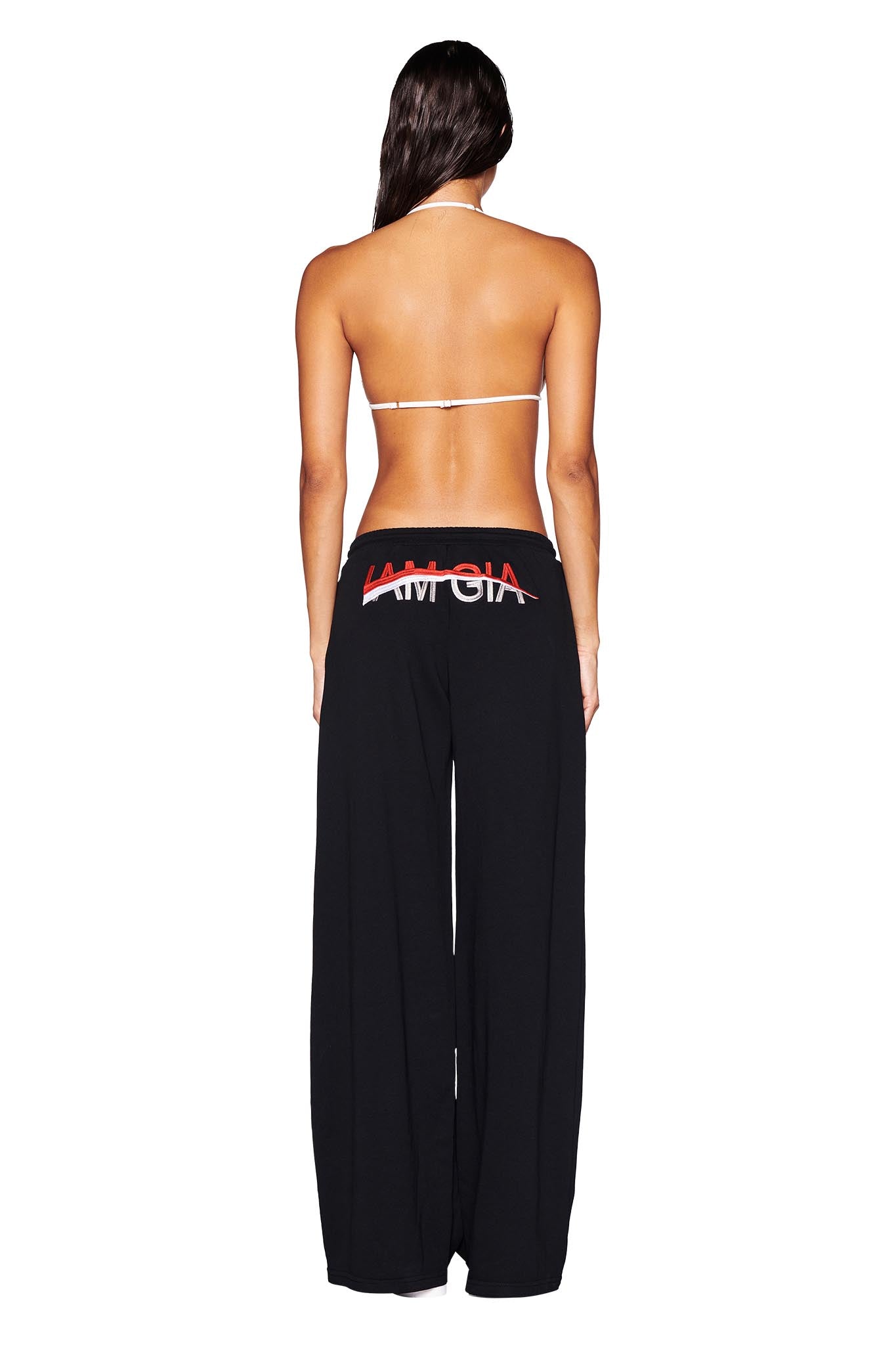 The image showcases a person from behind wearing a bikini top and the JUSTINE PANT - BLACK by I.AM.GIA. These black baggy track pants have a low-rise waist and feature the word "AMGIA" crossed out with a red line, set against a plain white background that accentuates the striking design.