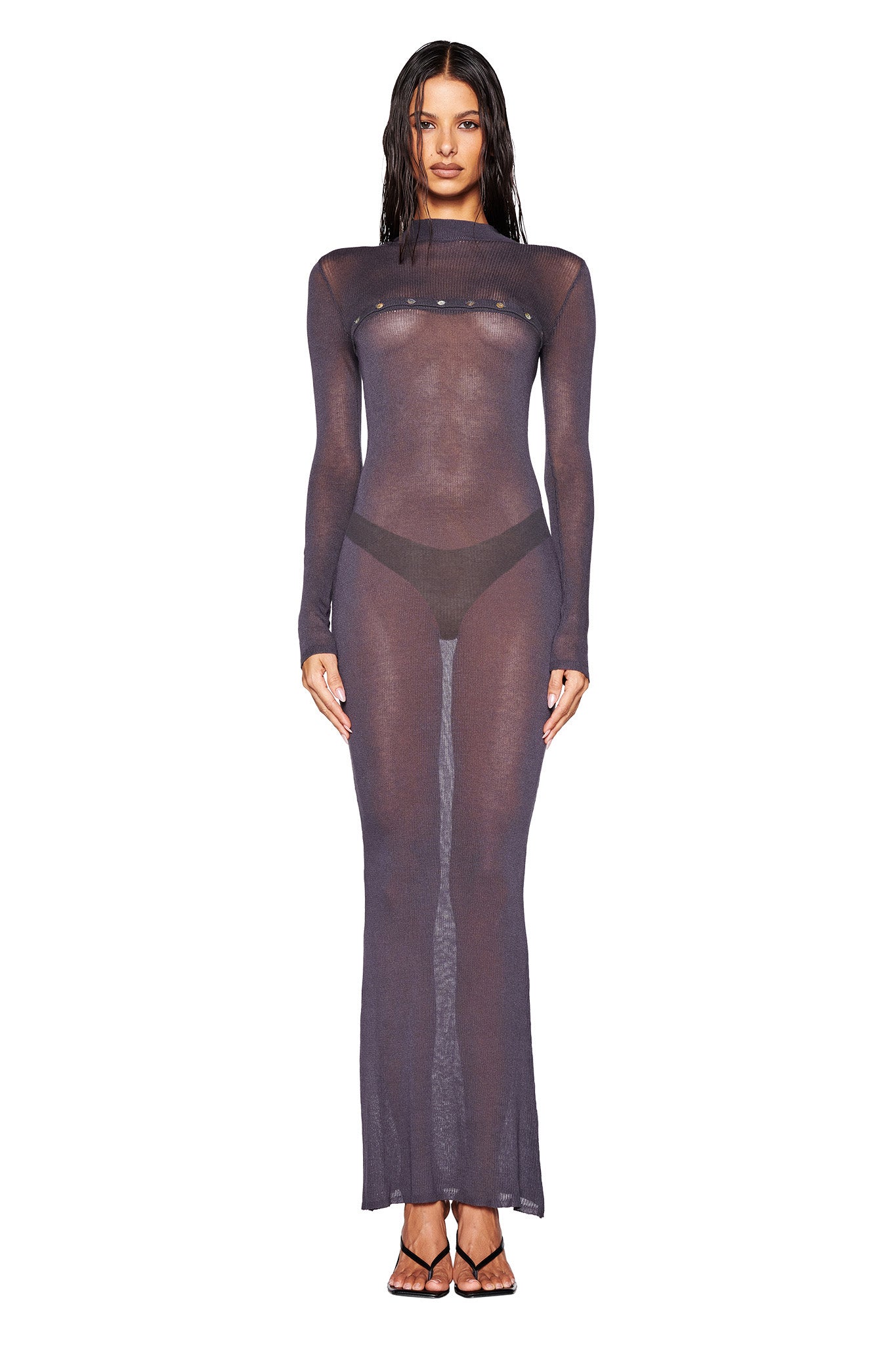 A person stands facing forward, dressed in the STORM MAXI DRESS by I.AM.GIA, featuring a long, sheer charcoal design layered over a black bikini and highlighting a mermaid silhouette. Their hair is long and wet, and they are wearing black strappy heels against a plain white background.