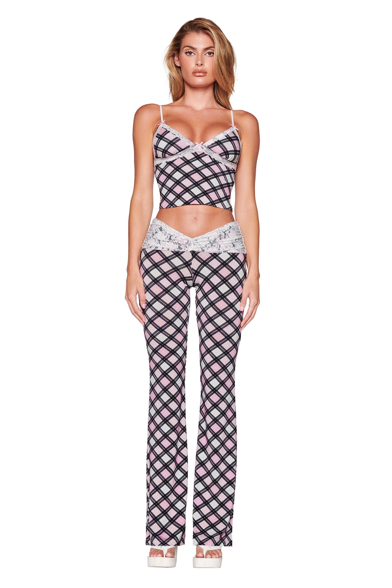 A person in a coordinating outfit by I.AM.GIA stands facing forward, wearing the KENDRA TANK - PINK in a plaid design, complemented by flared pants. This ensemble, characterized by adjustable shoulder straps, features a pink, white, and black geometric pattern. The look is completed with chic white platform sandals.