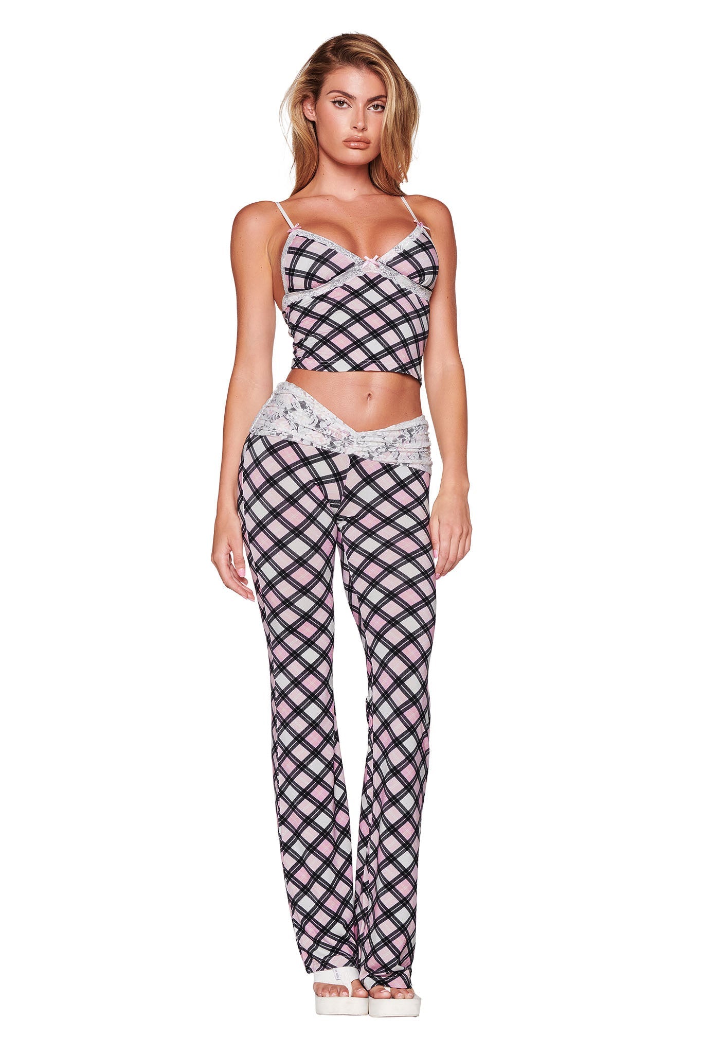 A person poses against a plain white background, wearing the KENDRA PANT - PINK : PLAID from I.AM.GIA, a matching plaid outfit consisting of a sleeveless crop top and low-rise flared silhouette pants. The ensemble showcases a striking black, pink, and white checkered pattern.