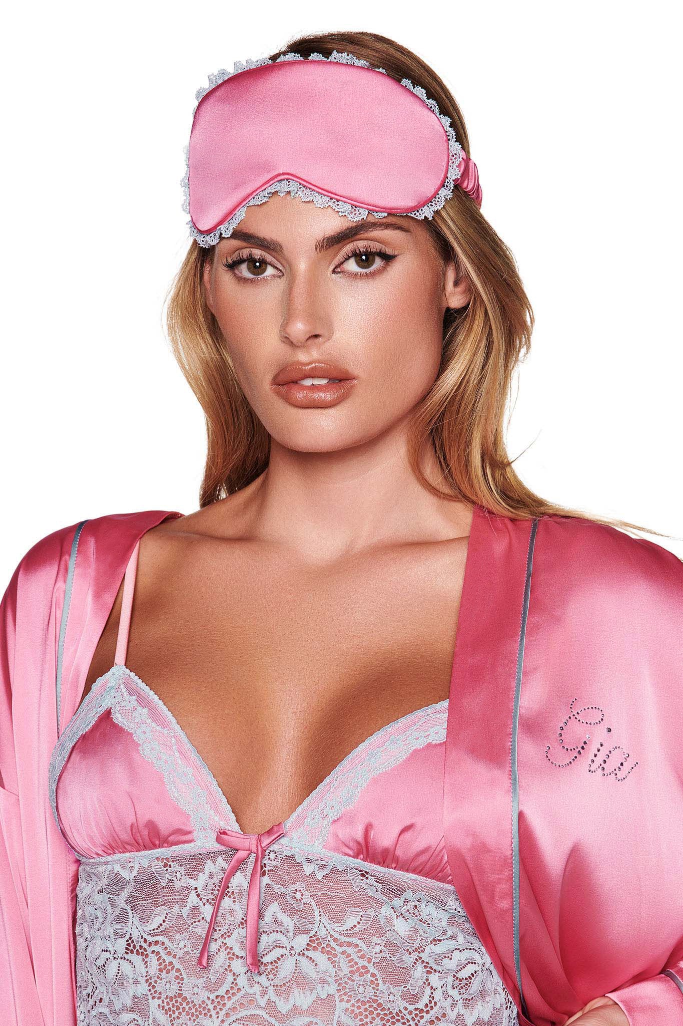 A person with long hair is wearing the I.AM.GIA MARTINA SLEEP ROBE in pink, featuring a relaxed fit with silky satin fabric and lace details. They complement the look with a matching pink sleep mask and a delicate lingerie top, all set against a plain white background.