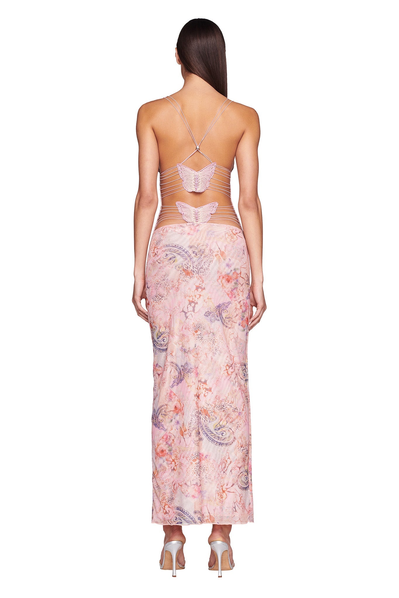 A woman with straight brown hair and heeled sandals faces away from the camera, wearing the I.AM.GIA ELLERY DRESS in Pink Flora Paisley—a long, sleeveless dress with an open back and thin crisscross straps.