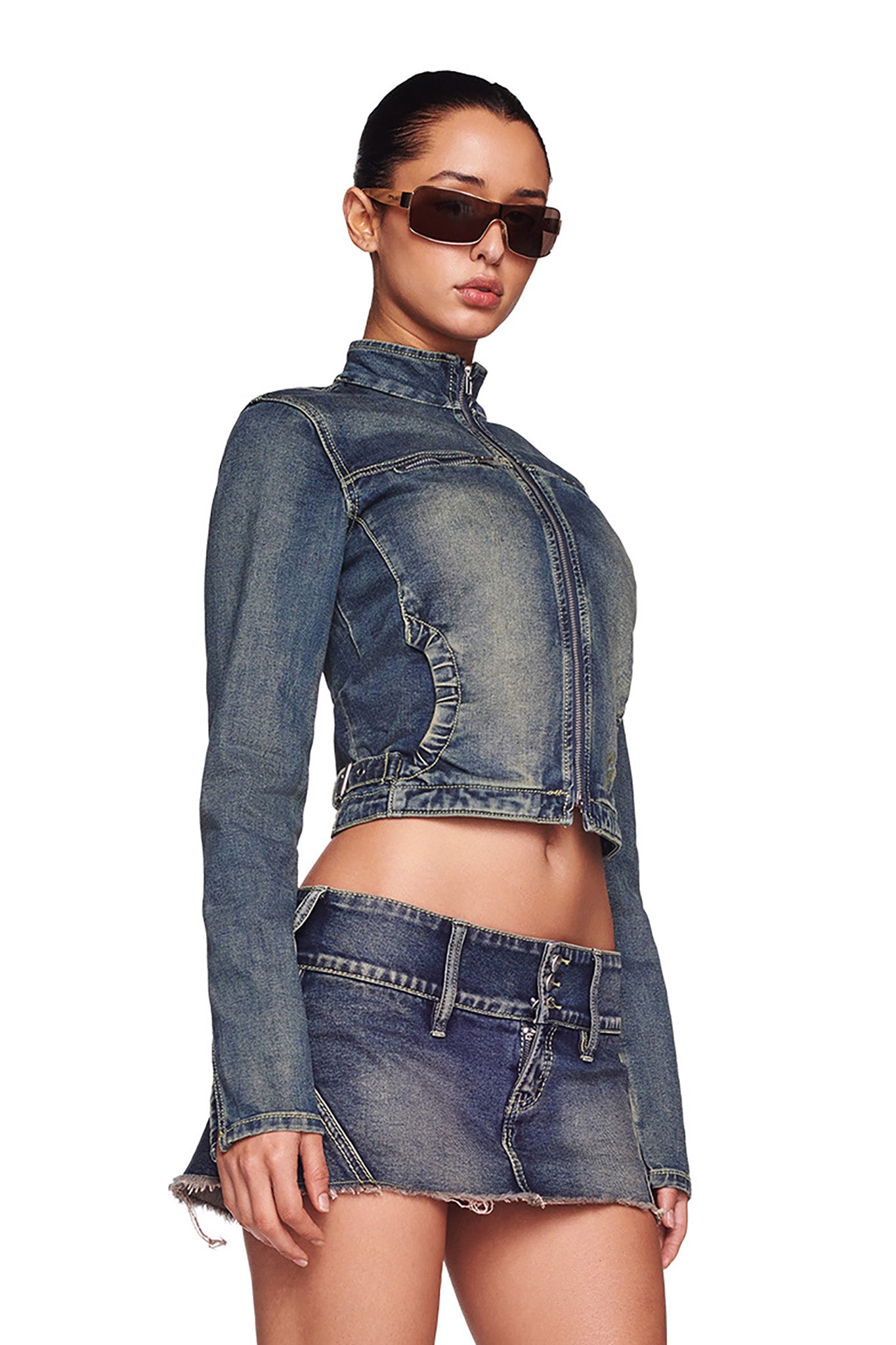 A person is wearing dark sunglasses, a fitted NYKKO JACKET in denim blue with long sleeves, and a matching I.AM.GIA mini skirt. Their hair is pulled back tightly. The outfit features distressed detailing on the jacket and skirt edges, set against a plain white background.