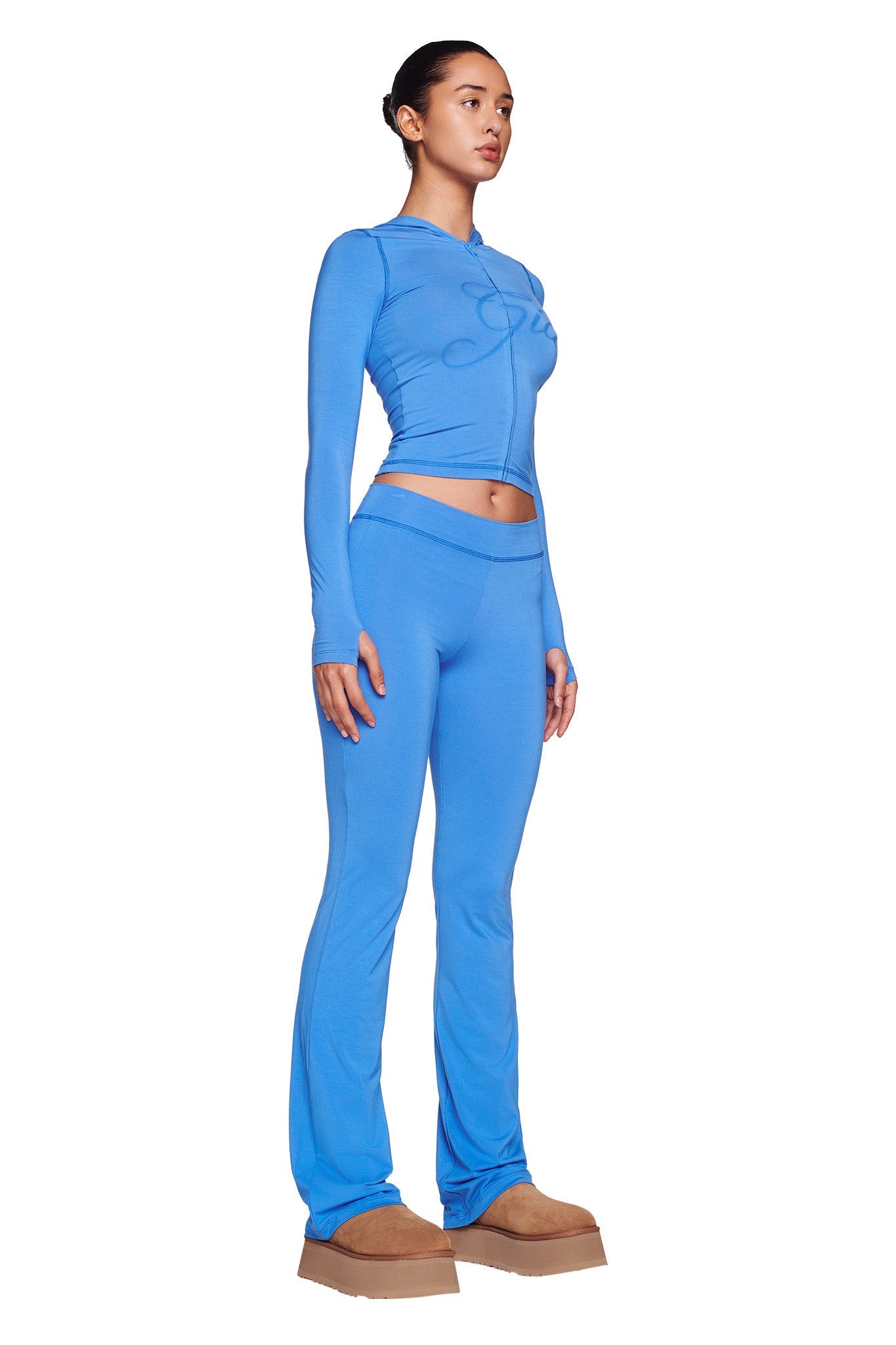 A woman stands against a white background, wearing a bright blue I.AM.GIA BLARE HOODIE with matching high-waisted, flared pants. She has brown hair styled in a sleek bun and is wearing light brown platform shoes. She gazes to the side with a calm expression.