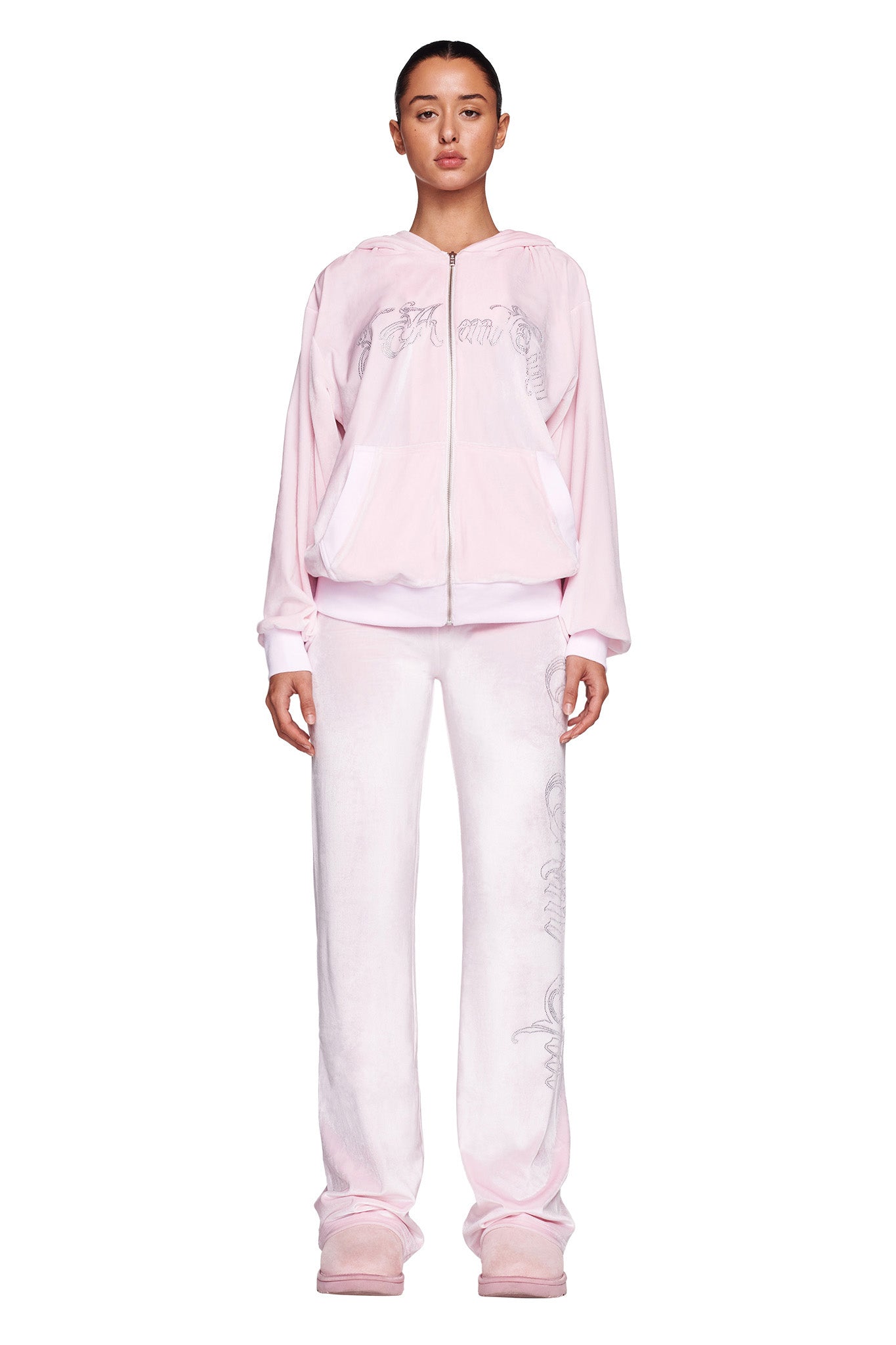 A woman stands against a white background wearing an I.AM.GIA light pink zip-up hoodie with a subtle graphic design, matching I.AM.GIA DARIA PANTS in ICY PINK, and light pink boots. Her hair is tied back, and she has a neutral expression.