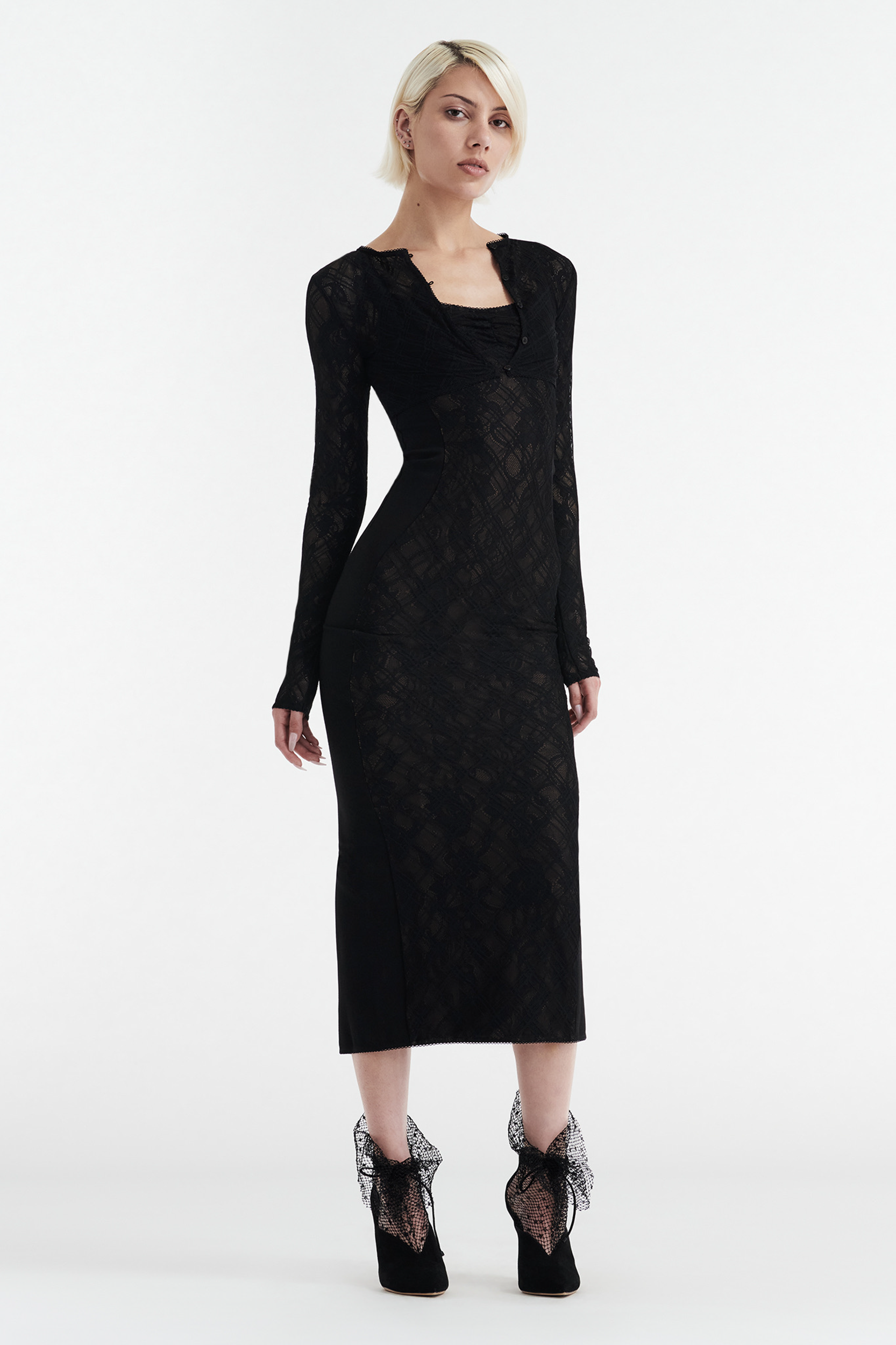A blonde woman models the CDP MARGOT TWO PIECE FITTED LACE MIDI DRESS in black, featuring long sleeves and elegant lace details, paired with black lace-adorned heels, as she stands confidently against a white background.
