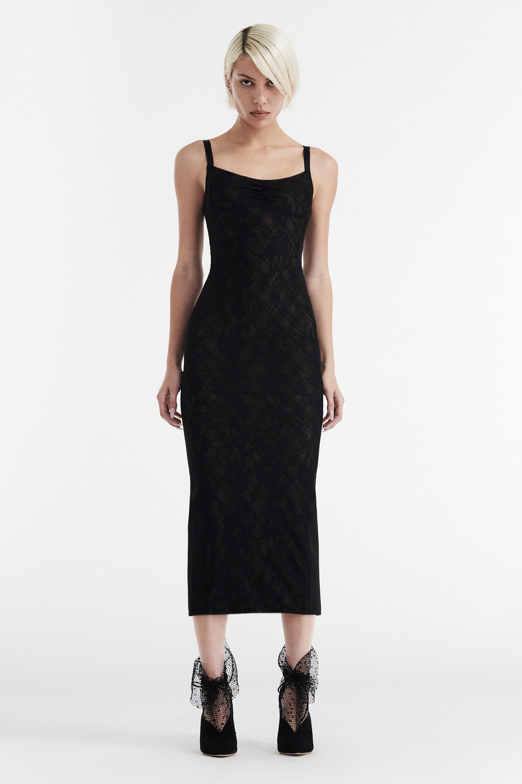 A woman with short blonde hair models the CDP MARGOT Two Piece Fitted Lace Midi Dress in black with black high heels featuring sheer bows, standing against a plain white background.