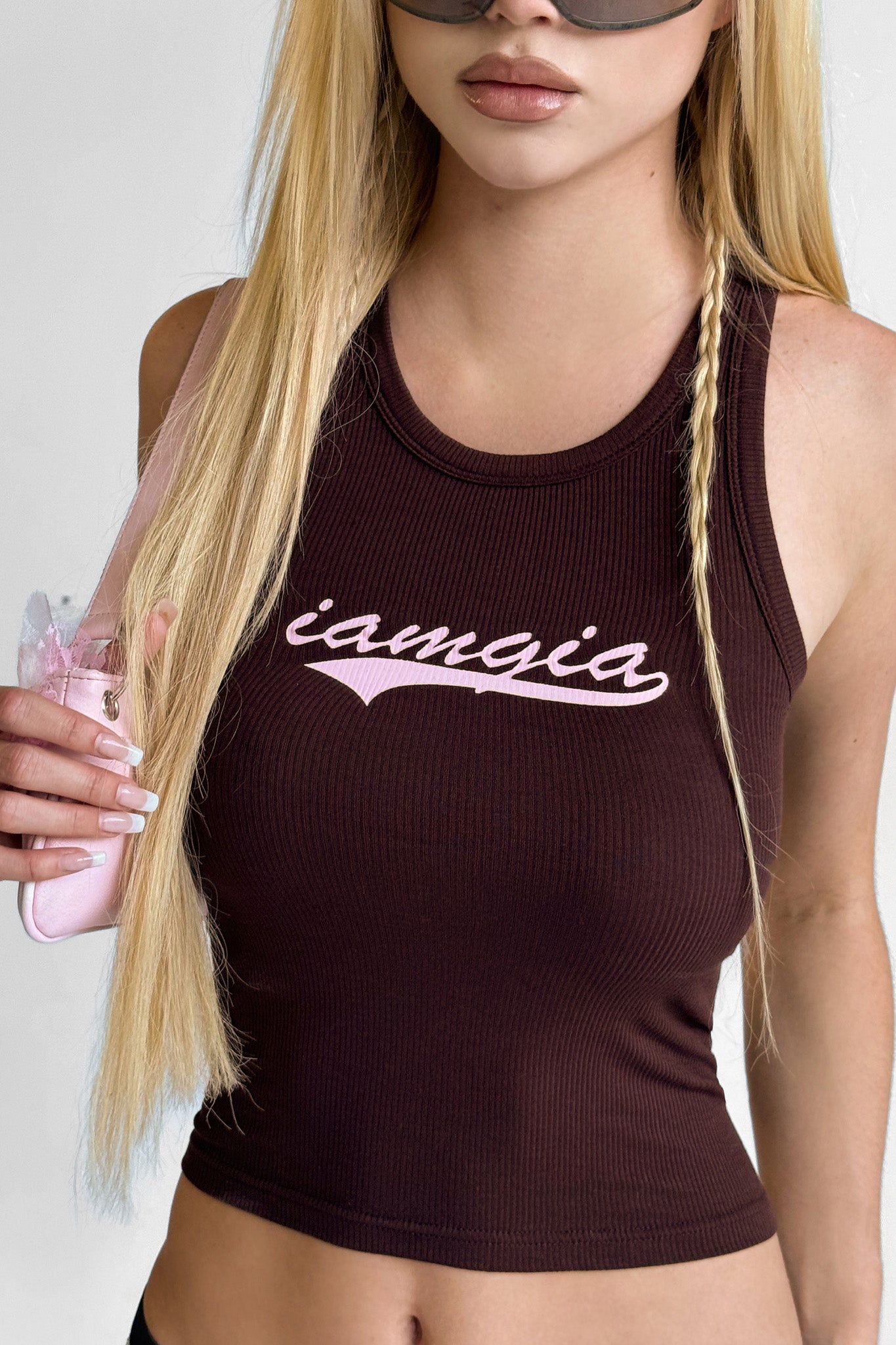 A woman with long blonde hair and sunglasses wears a sleeveless ribbed I.AM.GIA PENELOPE TANK in brown with "iamgia" in pink script, holding a light pink handbag over her shoulder.