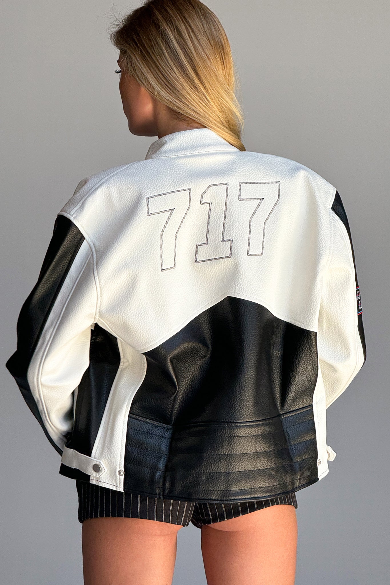 A person with long blonde hair is standing with their back to the camera, wearing an oversized silhouette I.AM.GIA ROXY JACKET in white and black, featuring the number "717" embroidered on the back. They are also sporting black pinstripe shorts. The background is plain and gray.