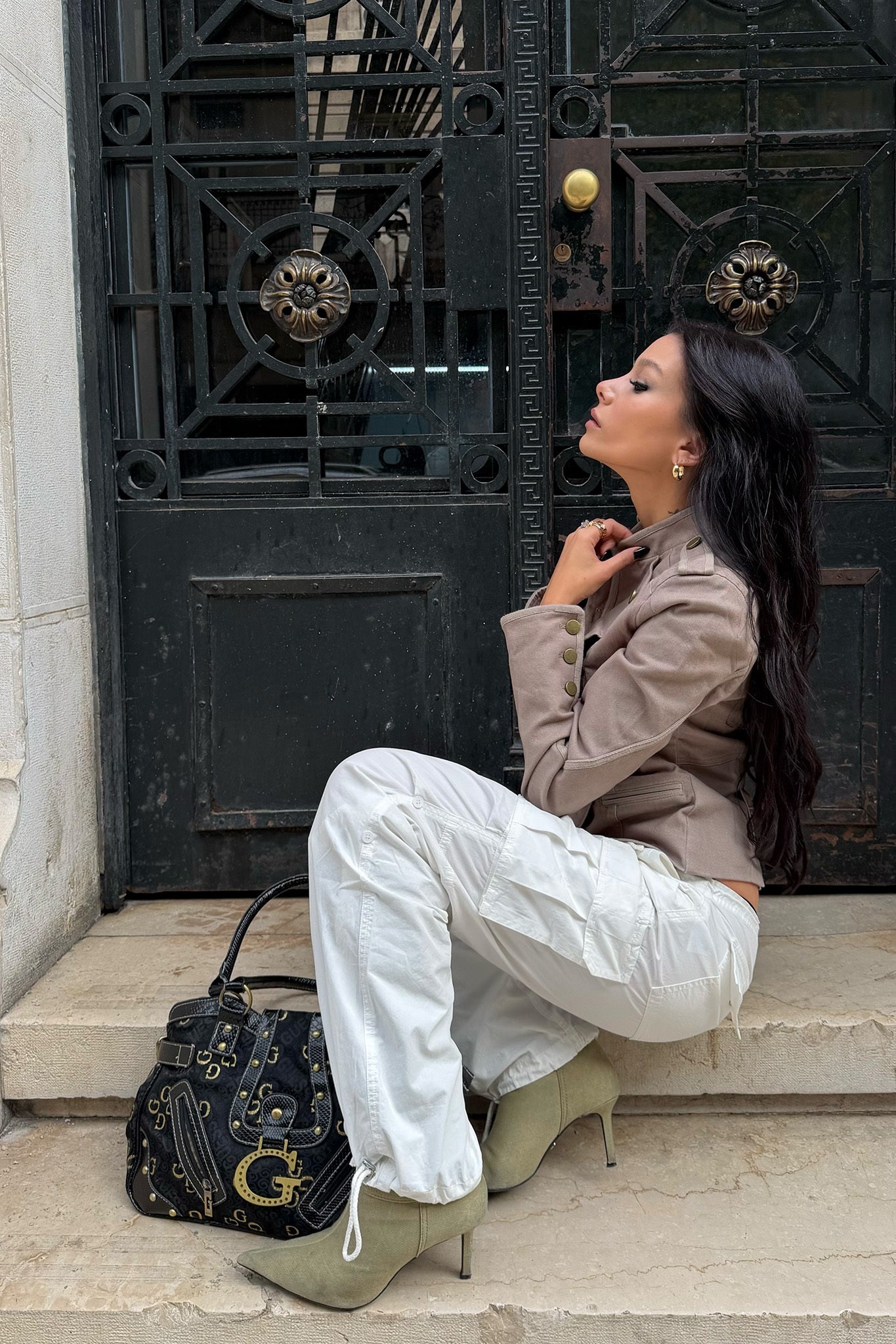 A woman with long dark hair sits on stone steps before a black door, wearing a beige jacket, green heeled boots, and the I.AM.GIA IVANA CARGO PANT in white. A black patterned handbag rests beside her.