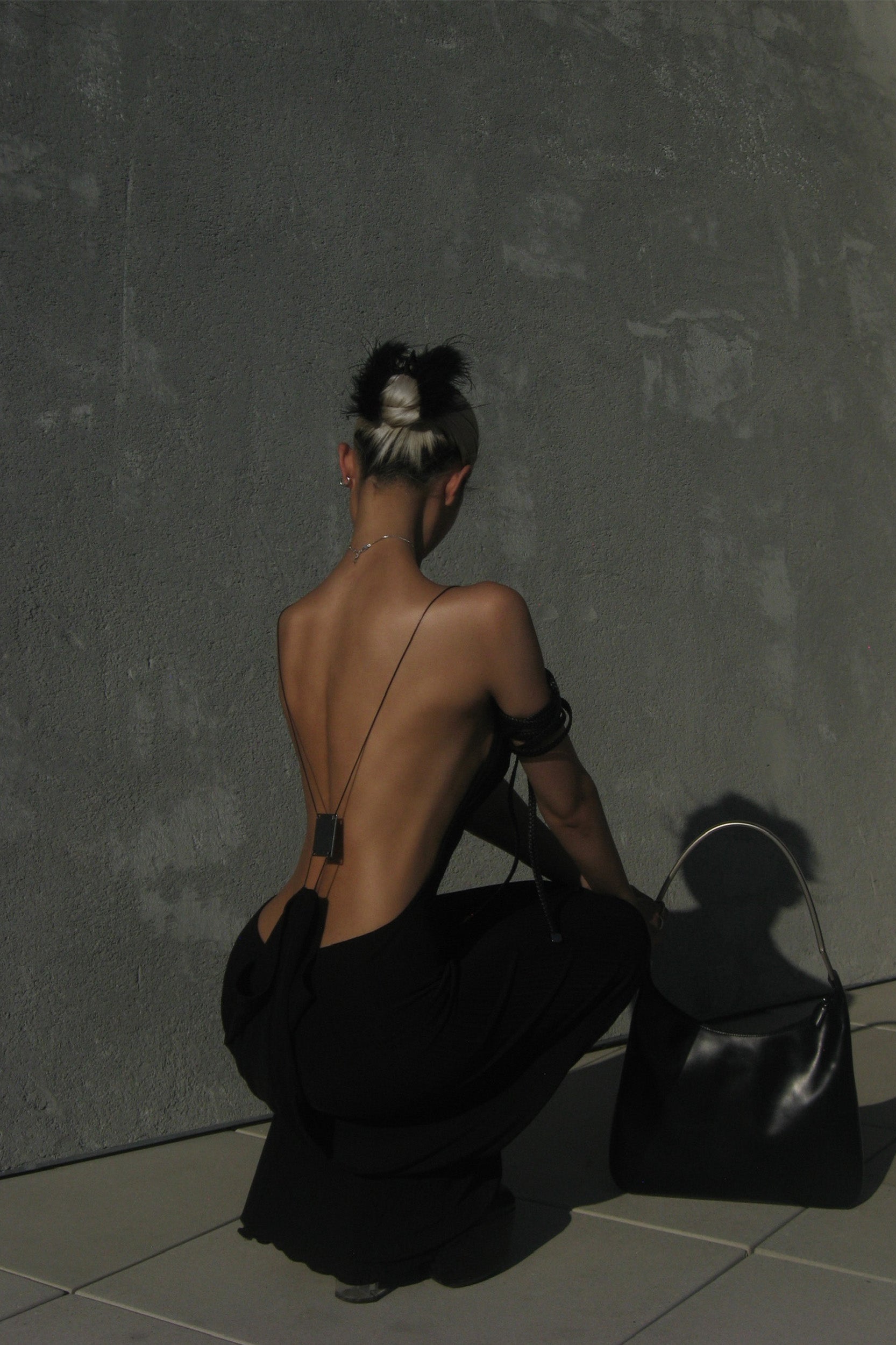 A person with a black and blonde hairstyle sits with their back to the camera, wearing the I.AM.GIA SIMONE MAXI DRESS in black. A large black handbag rests nearby against a textured gray wall.