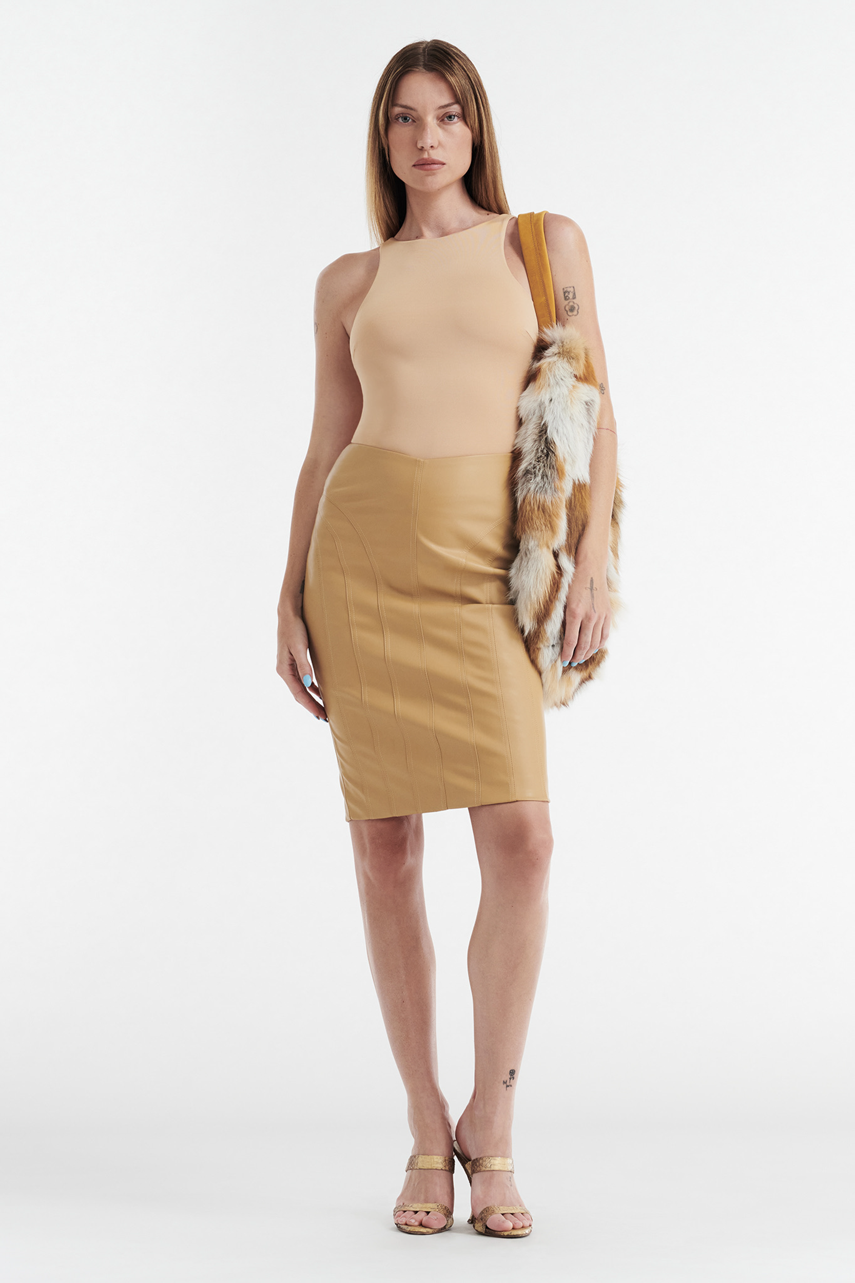 A woman faces forward in a CDP SOPHIE BASIC SLEEVELESS TANK WITH SHELF BRA in tan, paired with a tan knee-length skirt, heeled sandals, and a faux fur stole over one shoulder, set against a plain white background.