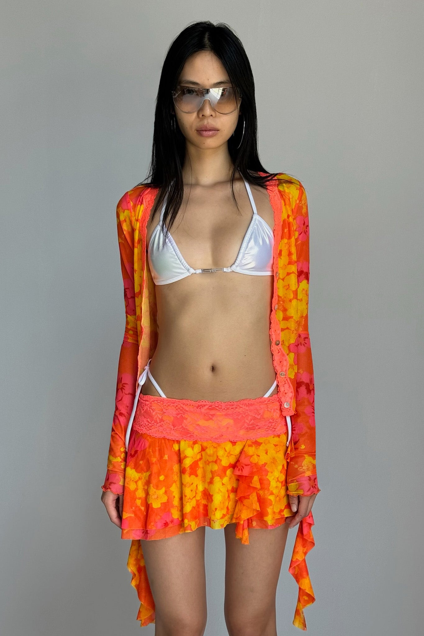 A woman with long dark hair models a sheer, colorful orange and yellow tie-dye I.AM.GIA cardigan over a white bikini top. She pairs it with matching ruffled mini skirt, the ALOHI BIKINI BOTTOM - WHITE : PEARL from I.AM.GIA, and oversized sunglasses. She stands against a plain gray background, gazing directly at the camera.