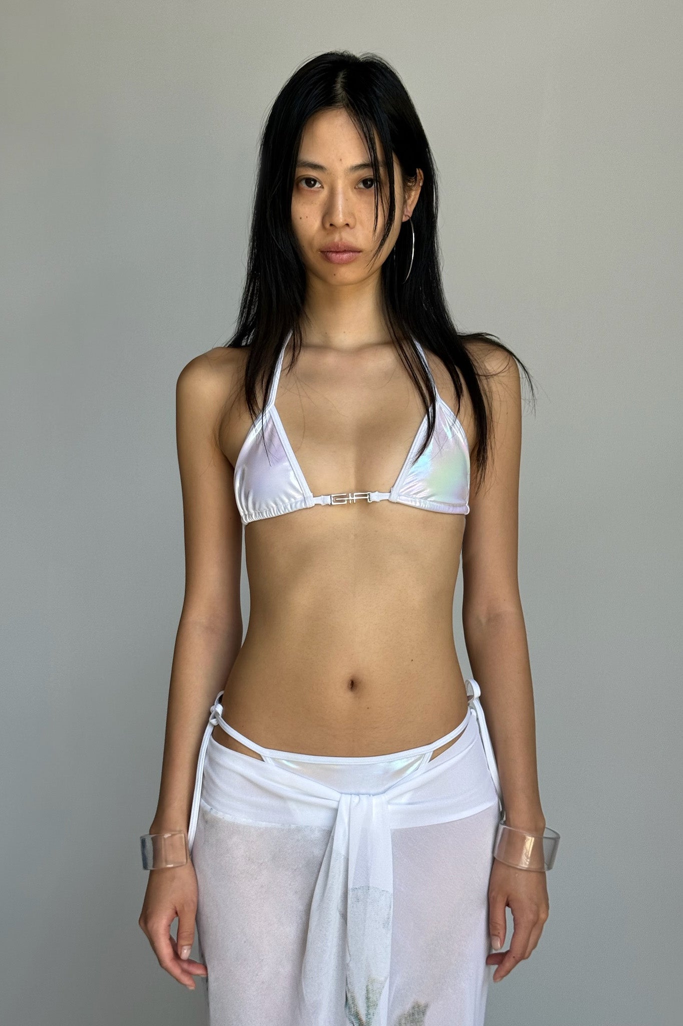 A woman with long black hair, dressed in the I.AM.GIA ALOHI BIKINI TOP - WHITE : PEARL and a sheer white sarong, stands against a plain grey background. She wears transparent bracelets on both wrists and looks directly at the camera.