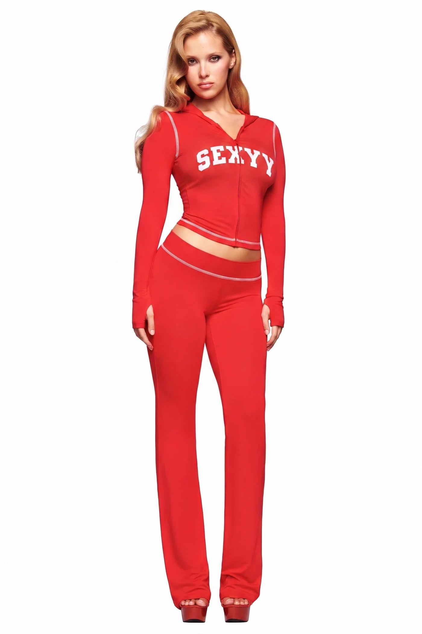A woman faces forward in an I.AM.GIA X SEXYY RED BLARE TRACKPANT set, featuring a fitted red zip-up jacket with "SEXYY" printed on it and matching low-rise pants, set against a white background.