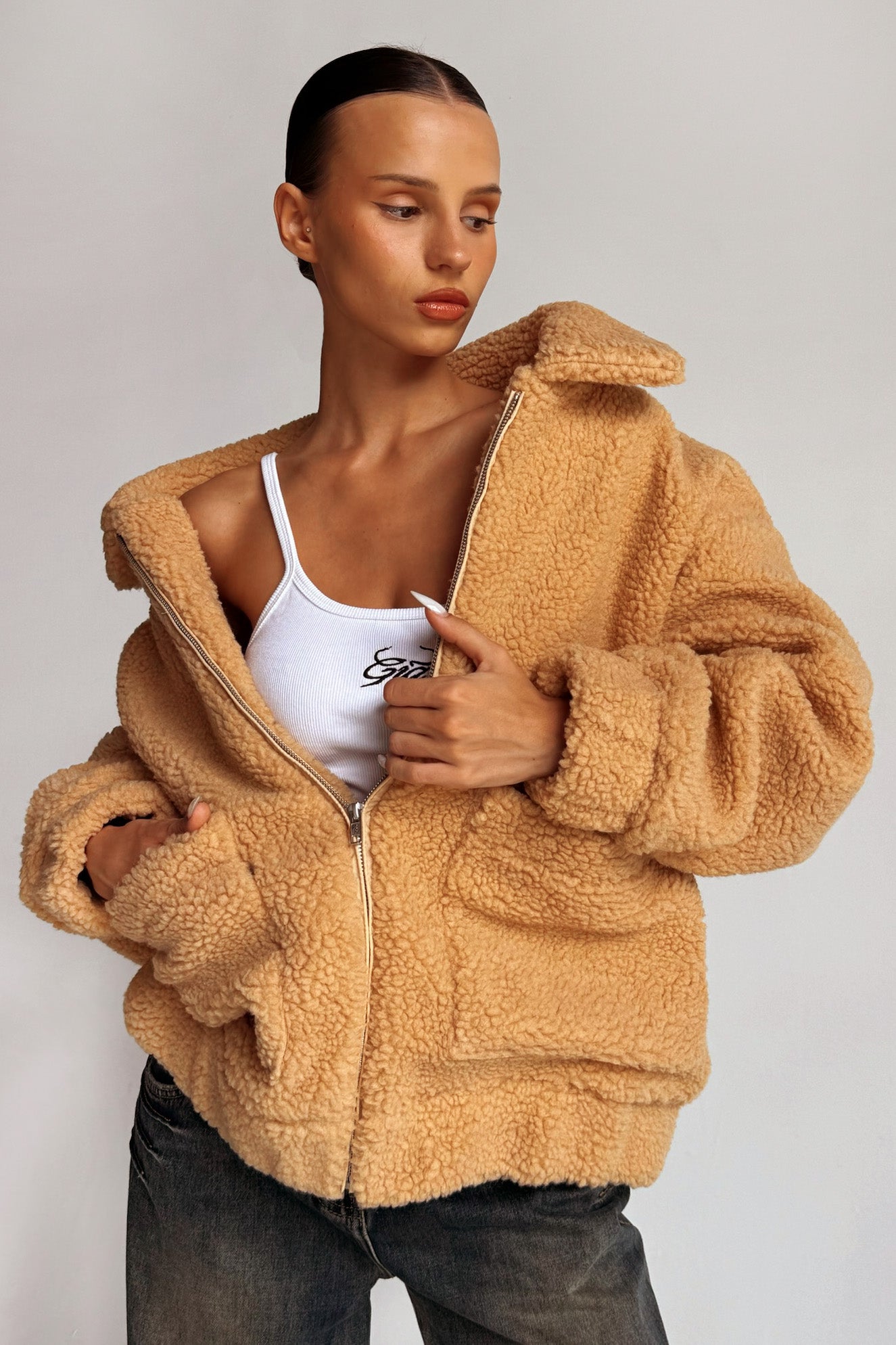 A person with slicked-back hair is wearing the PIXIE COAT - CARAMEL from I.AM.GIA over a white tank top with text on it. They have an intense expression and are lightly gripping the coat’s zipper with one hand. The background is plain and gray.