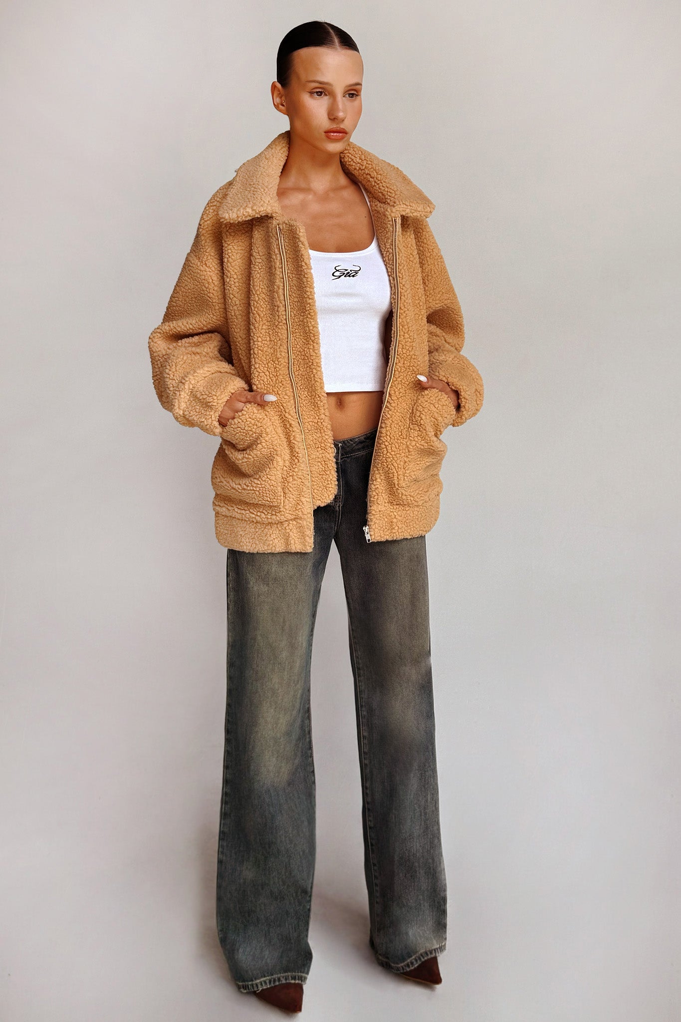 A woman stands against a plain background wearing the PIXIE COAT - CARAMEL from I.AM.GIA over a white crop top and loose-fitting gray jeans. She has her hands in the coat pockets and is looking slightly to the side, showcasing a casual, yet stylish look.