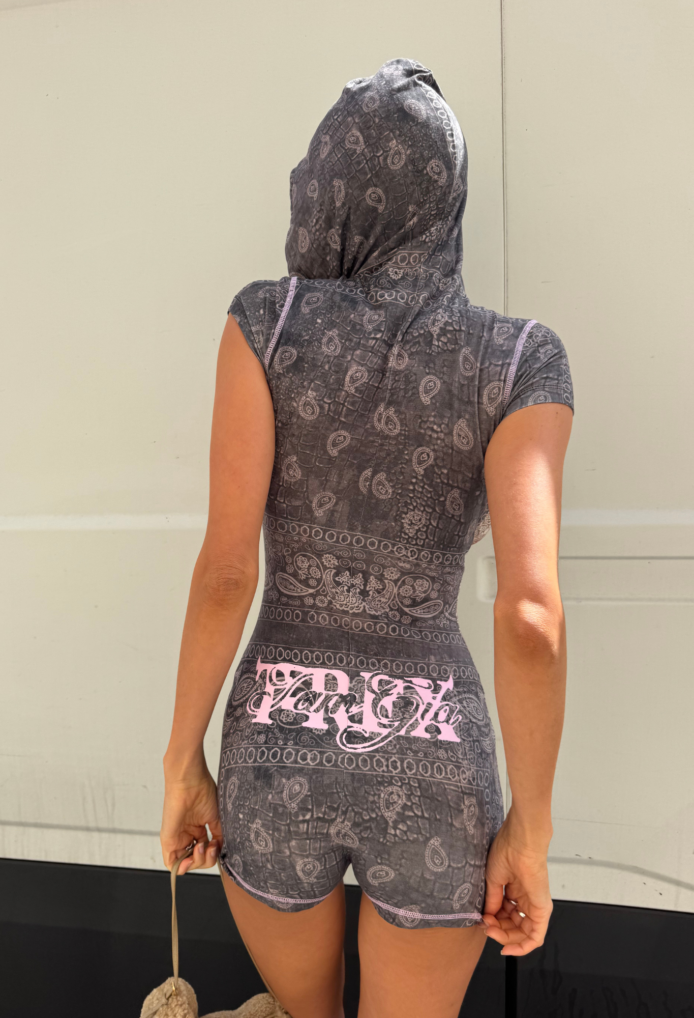 A person stands facing away, wearing the I.AM.GIA T-REX BLARE Short Sleeve Romper in Charcoal Bandana—a hooded romper showcasing the brand’s exclusive gray and pink bandana print. They hold a rope bag, set against a light-colored wall with a black stripe at the bottom—making this outfit an ideal festival statement.