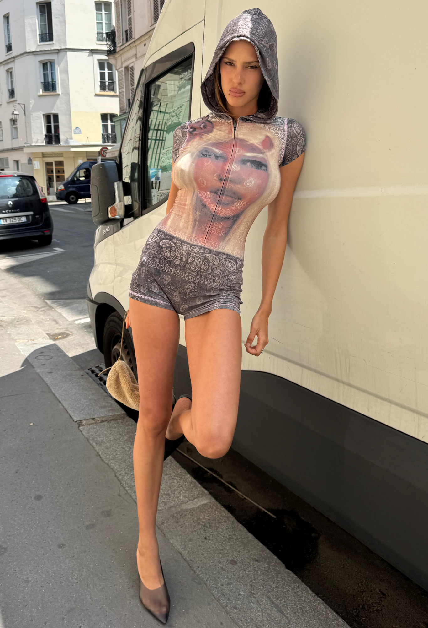 A person wears the I.AM.GIA T-REX BLARE SHORT SLEEVE ROMPER in Charcoal Bandana, leaning against a white van on a city street with one leg bent and holding a small tan purse—a bold festival statement outfit.