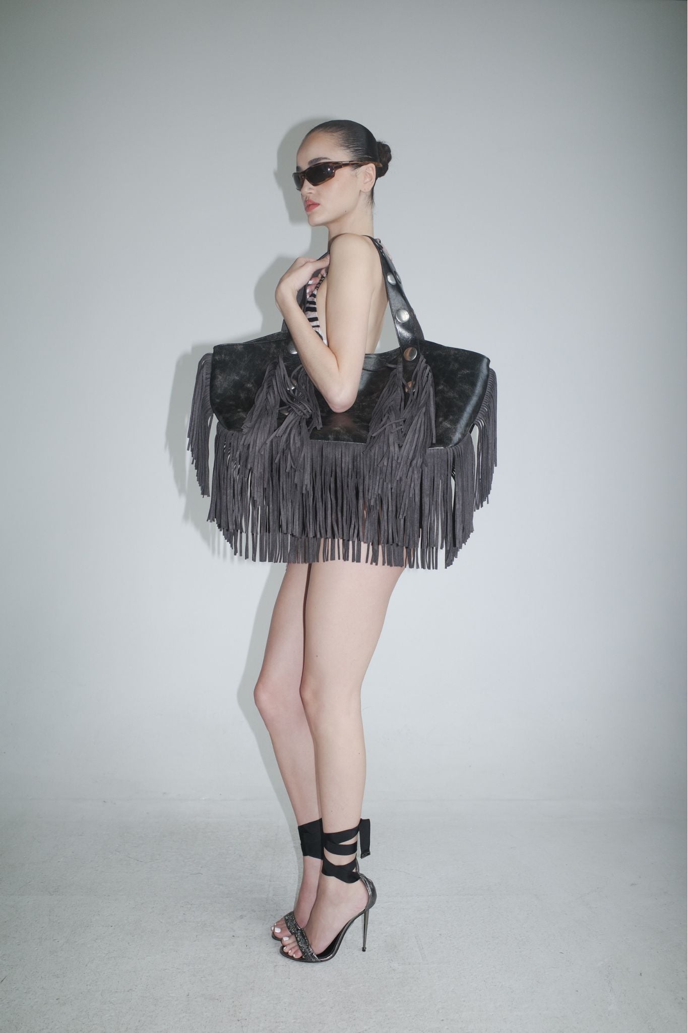 A woman in sunglasses, high heels, and a swimsuit poses by a plain wall, carrying the I.AM.GIA CHROMA FRINGE BAG - BLACK with distressed black fringe on her shoulder.