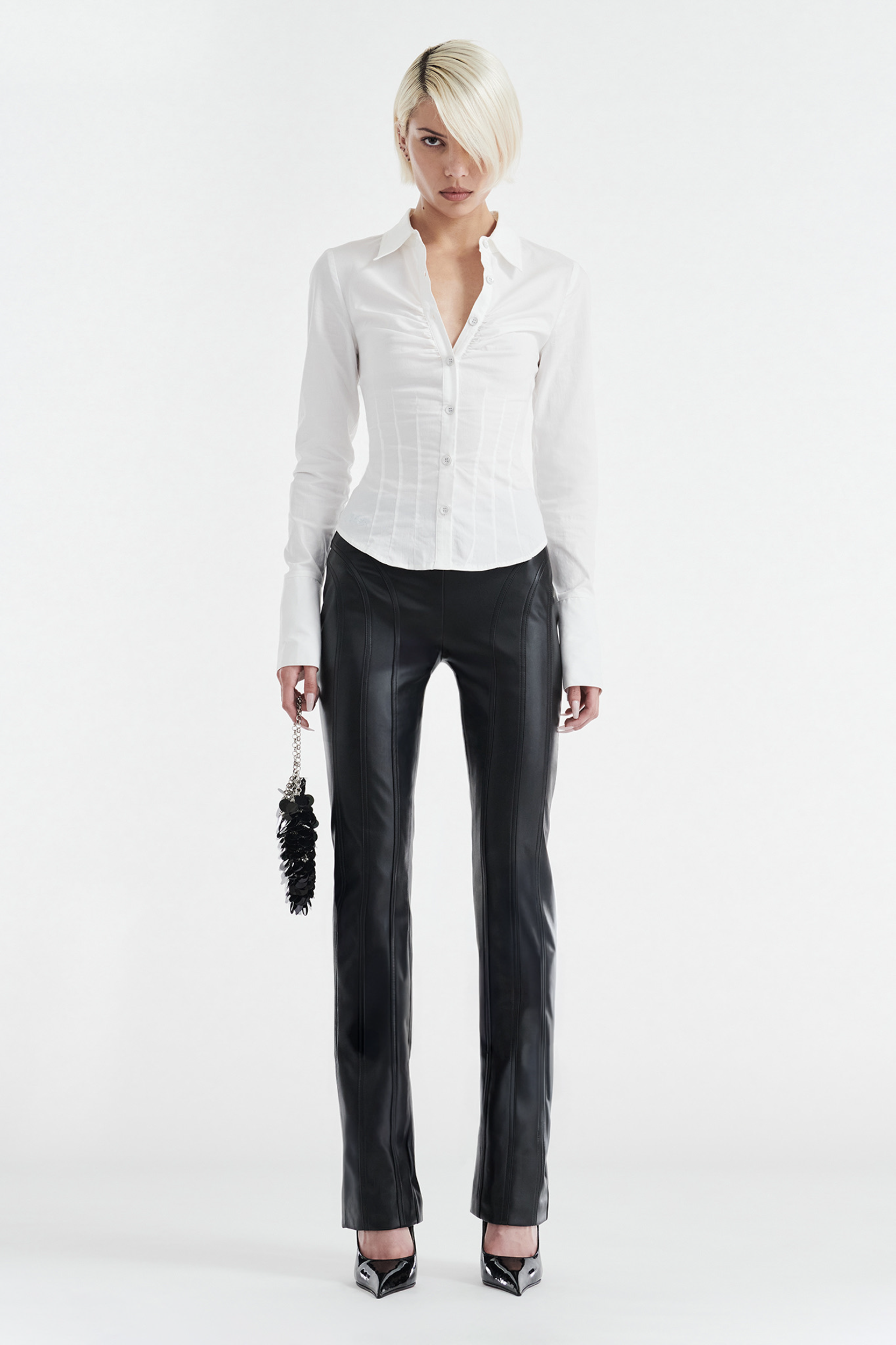 A person with short blonde hair wears the CDP ZOE LONG SLEEVE COTTON BUTTON UP SHIRT in white, black leather pants, and black pointed heels, holding a small black accessory in their left hand against a plain white background.