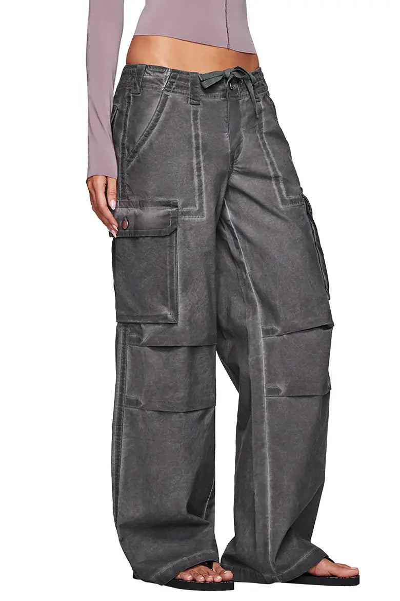 A person models the I.AM.GIA JAXXON CARGO PANT in washed black, featuring a low rise, baggy leg, large pockets, and seam details—paired with a long-sleeve light mauve top and black sandals. The image focuses on the pants from waist to feet.