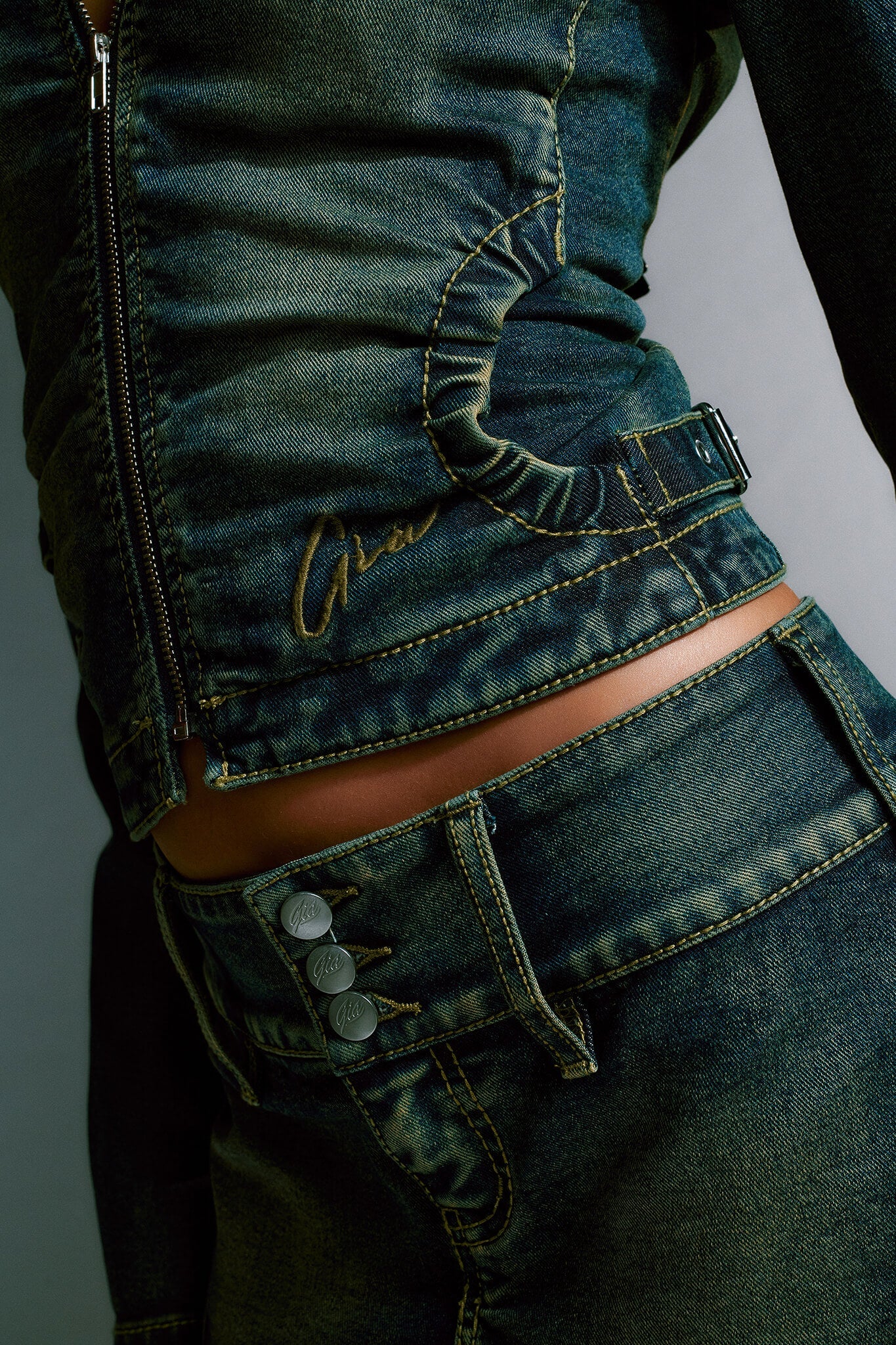 Close-up of a person wearing the stylish, fitted NYKKO JACKET - BLUE from I.AM.GIA, paired with matching high-waisted jeans. The outfit features detailed stitching, a zip closure on the jacket, and three metal buttons on the jeans. The dark wash denim has a slightly worn, vintage look.