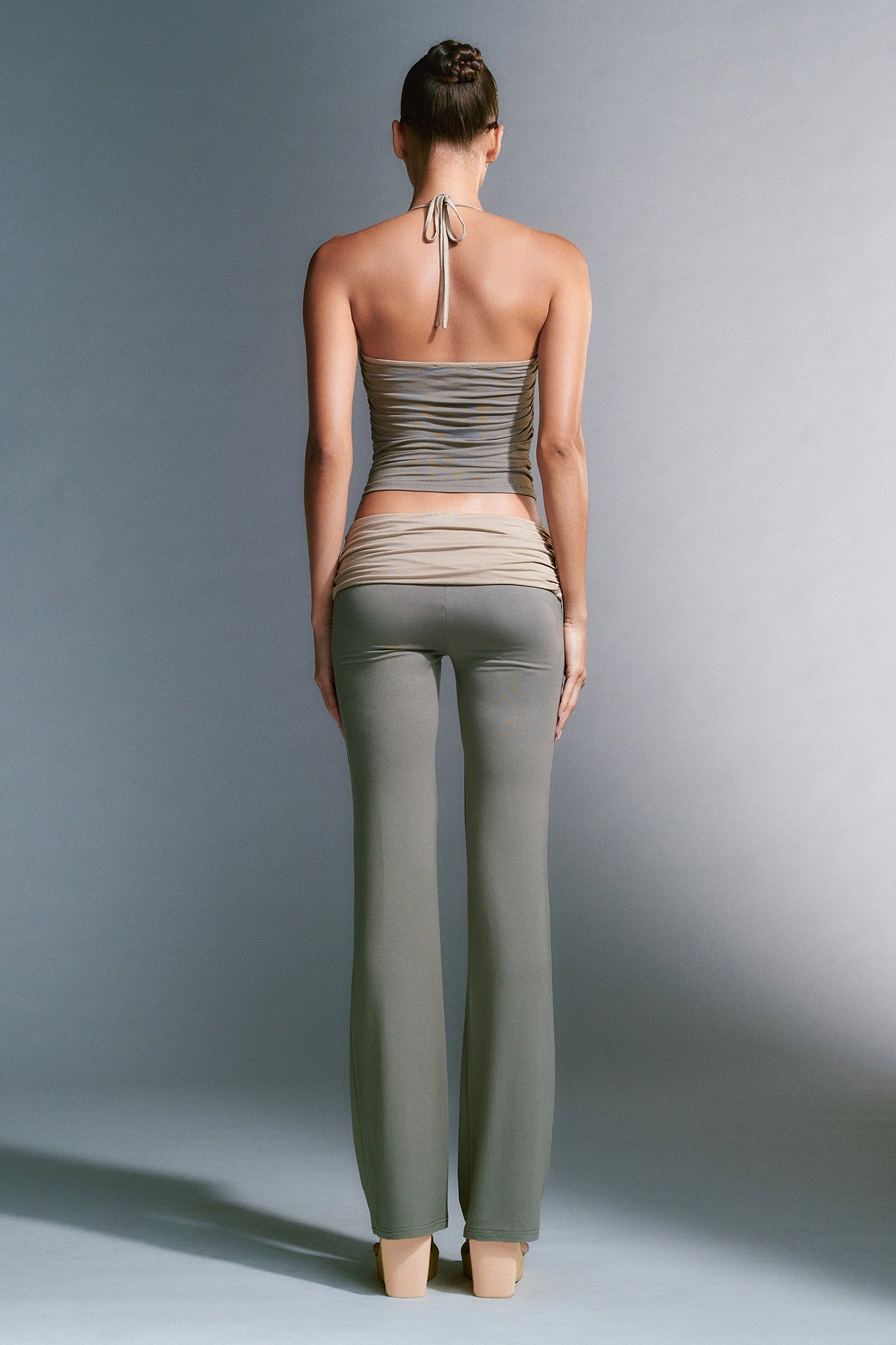 A person with tied-back hair stands facing away, wearing a backless, halter-neck, ribbed green tank top named JANNIS TANK by I.AM.GIA and matching high-waisted, flared grey pants. The background is a neutral grey. Their pose accentuates the slim fit of the clothing.