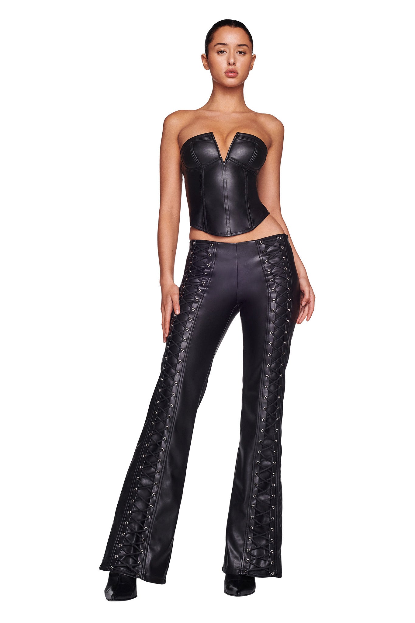 A woman is standing against a white background, wearing the I.AM.GIA EVANNA CORSET in black, paired with matching high-waisted leather pants featuring lace-up detailing along the sides. Her hair is pulled back as she poses with her hands slightly placed on her hips.