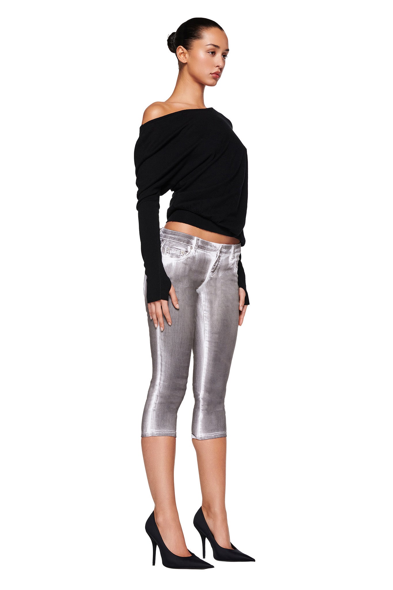 A woman stands in profile against a white background, wearing the GISELLE TOP - BLACK with long sleeves from I.AM.GIA, silver metallic cropped pants, and black high-heeled shoes. Her dark hair is styled in a bun.