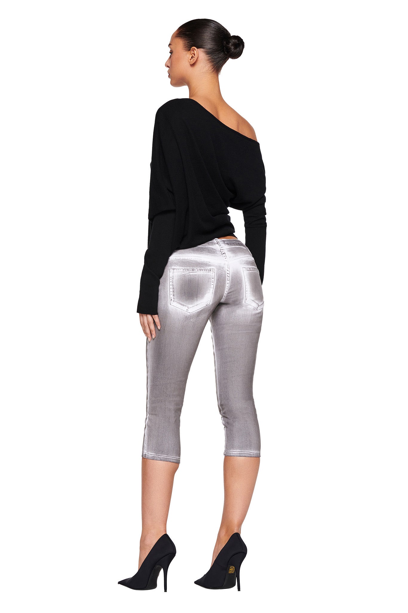 A woman stands with her back to the camera in a stylish pose. She has her dark hair in a neat bun and is wearing the GISELLE TOP - BLACK by I.AM.GIA, along with long sleeves, metallic silver capri pants also by I.AM.GIA, and black high heels.