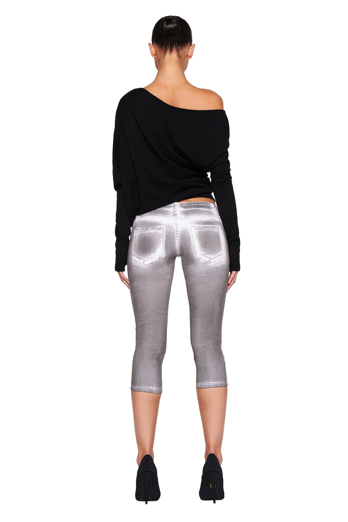 A person with short hair stands with their back to the camera, embodying I.AM.GIA style. They wear the GISELLE TOP - BLACK from I.AM.GIA, paired with shiny silver-gray capri pants and black high heels. The crisp white background accentuates their bold fashion statement.
