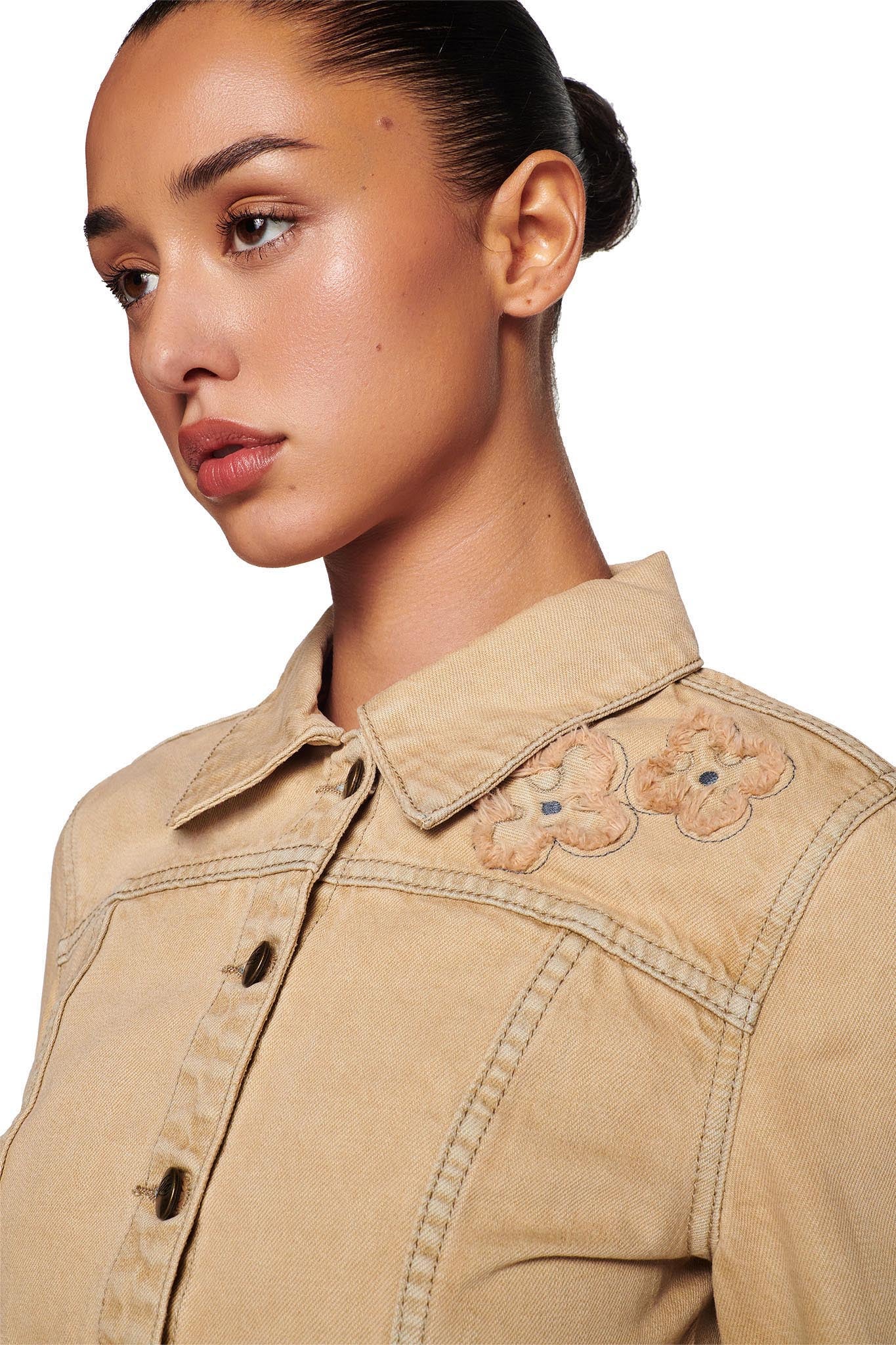 A young woman with her hair pulled back is wearing the FAUNA JACKET - BEIGE : TAN DENIM from I.AM.GIA, which features embroidered flowers on the shoulder. The image captures her in a side profile view against a plain white background.