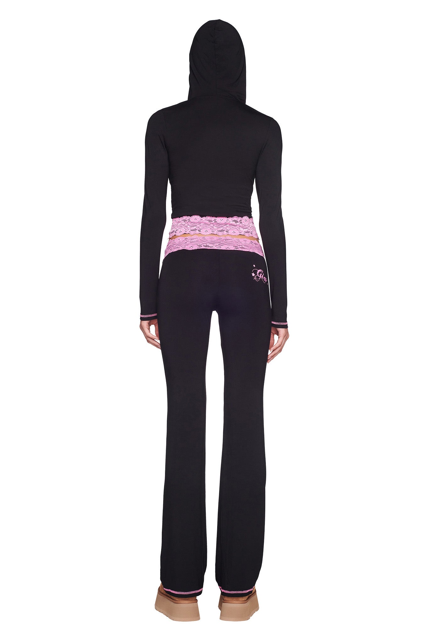 A person wearing the TARSHA JACKET in black/pink from I.AM.GIA, black flared pants with a pink lace waistband and a small "JS" design on the left hip, along with beige shoes stands facing away.