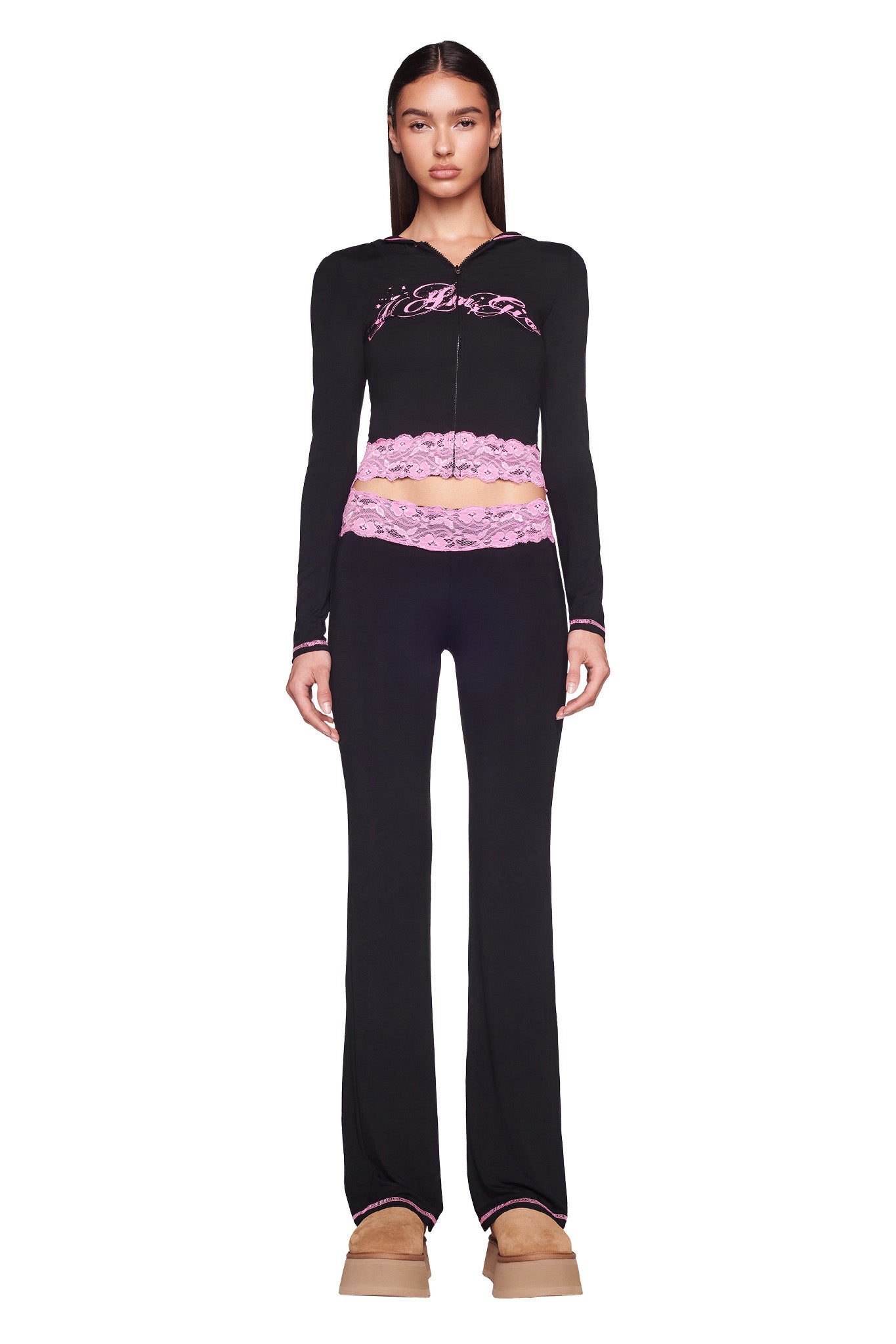 A woman is standing against a white background, wearing the I.AM.GIA TARSHA JACKET in black and pink. She pairs it with matching black pants and light brown platform shoes. Her straight hair is down, and she has a neutral expression.