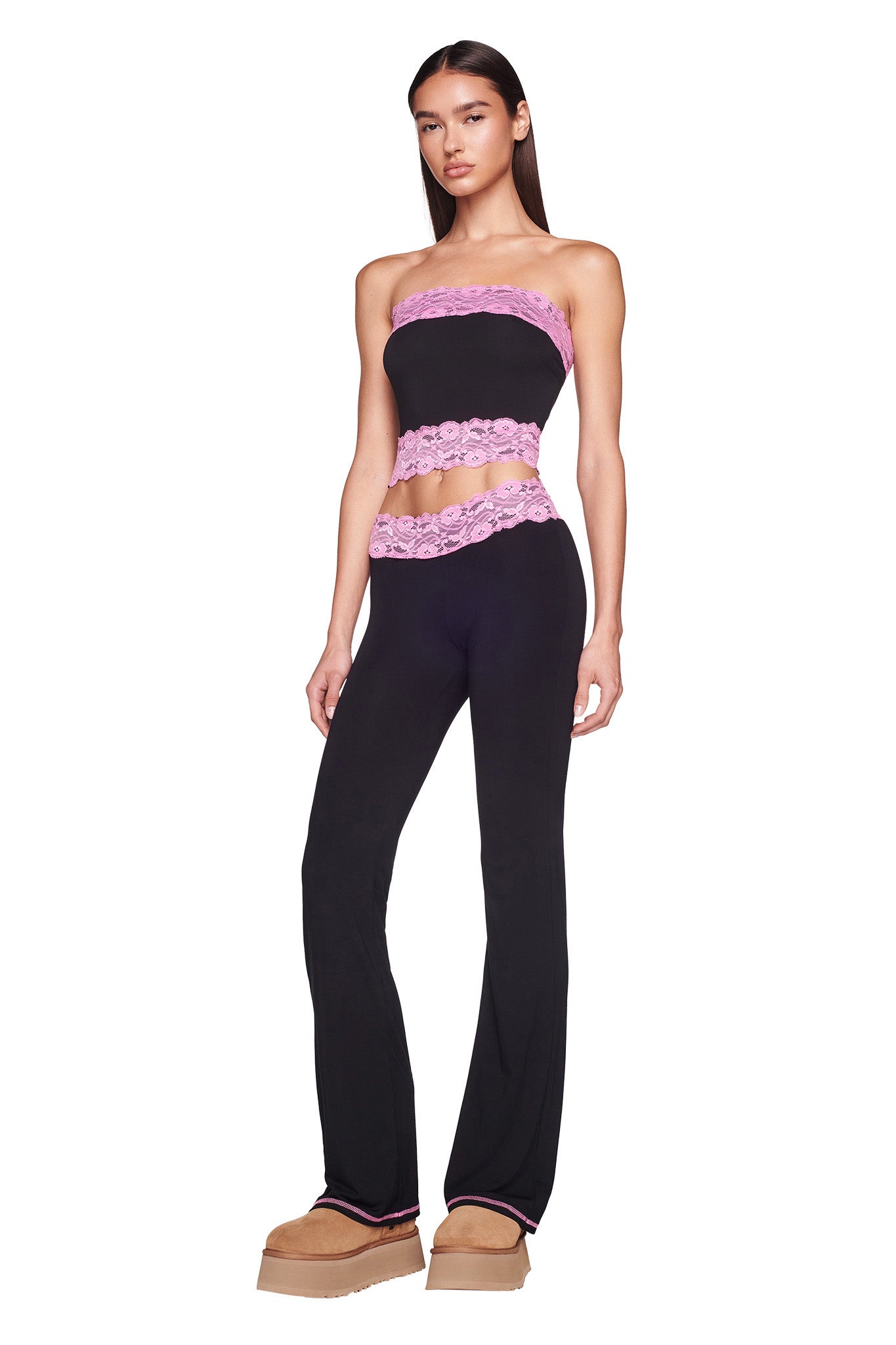 A woman with long dark hair stands facing forward, her left hand resting on her thigh. She wears the TARSHA PANT in black with pink lace trim from I.AM.GIA, paired with a matching strapless black top featuring similar pink lace detail. Her outfit is completed with light brown platform shoes.