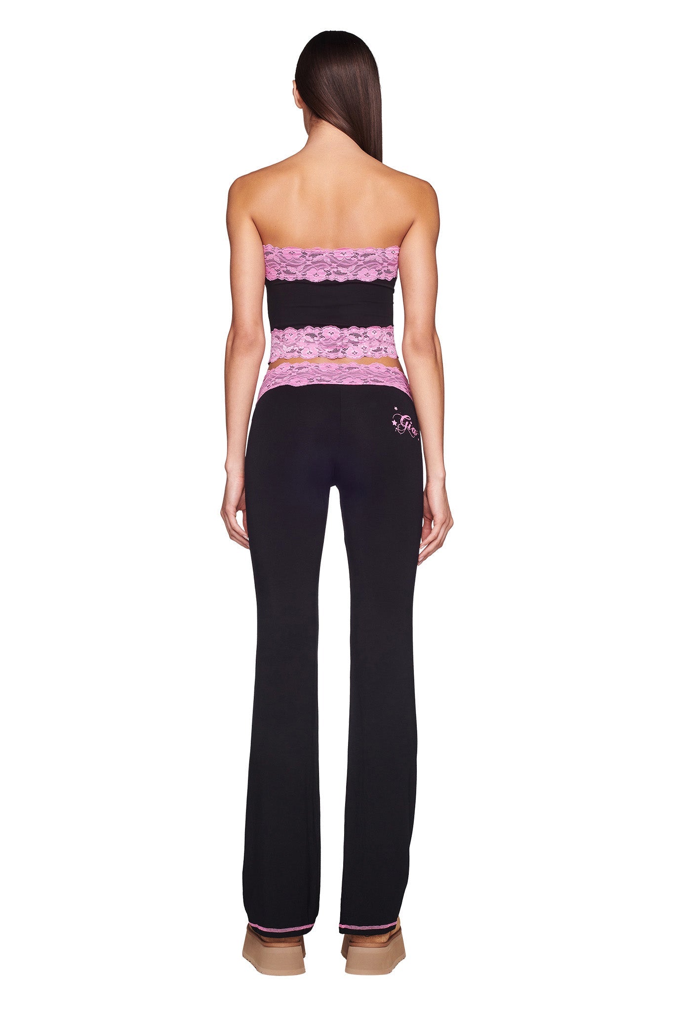 A woman with long, straight hair faces away from the camera, showcasing the I.AM.GIA TARSHA BOOB TUBE in black and pink, paired with matching pants featuring pink accents. Her platform shoes complete the look, while her bare arms highlight the form-fitting outfit.