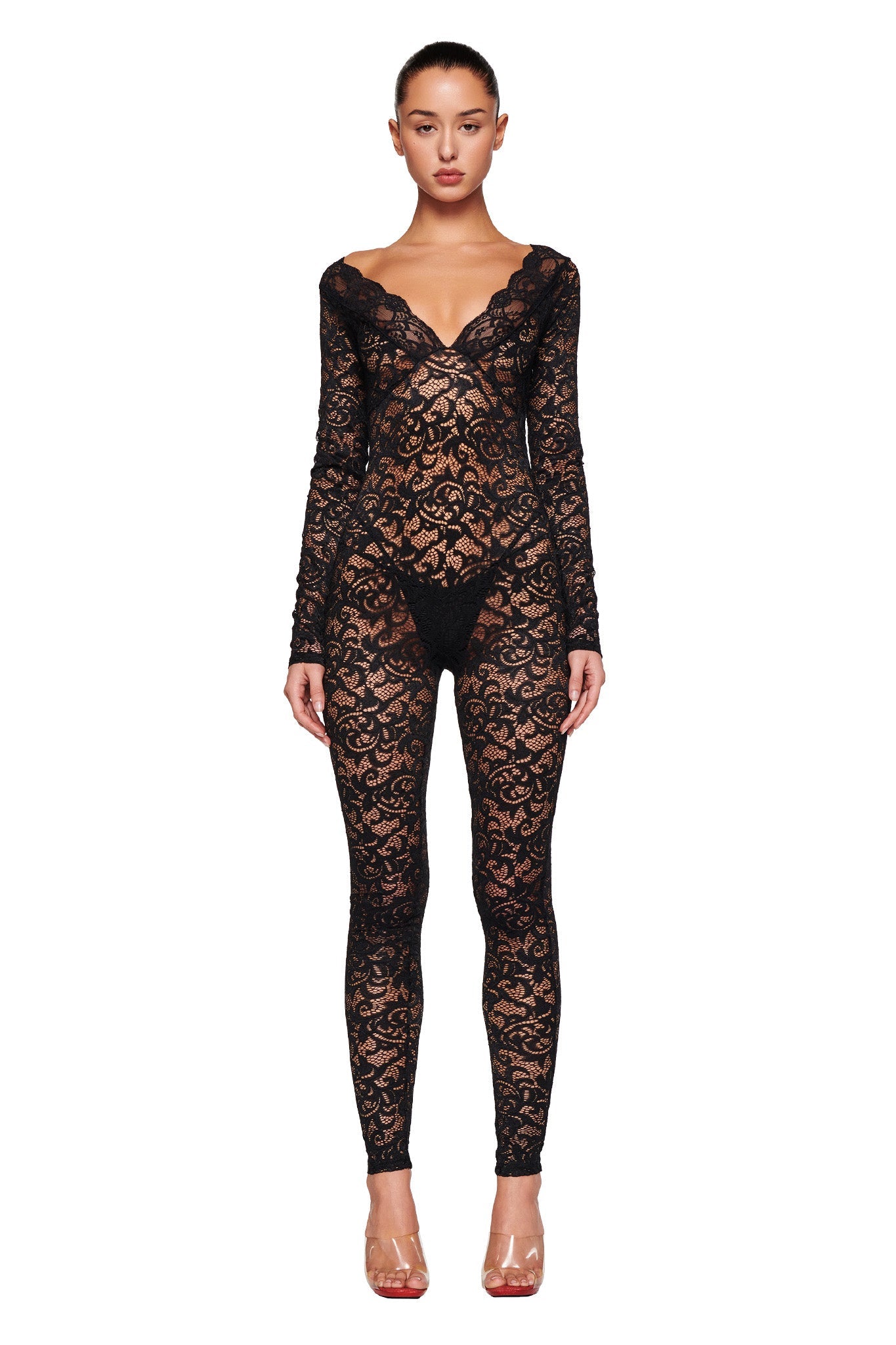 A woman stands facing the camera, wearing a full-length, long-sleeved GEMMA CATSUIT - BLACK by I.AM.GIA with a plunging neckline. The intricate lace pattern is sheer, and she pairs the outfit with clear high-heeled shoes. Her hair is styled in a sleek, pulled-back look.