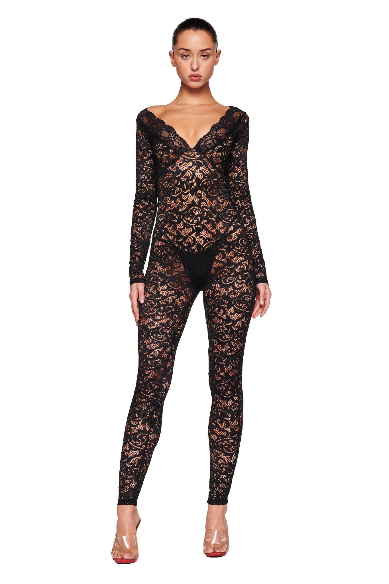 A model stands facing forward, wearing the GEMMA CATSUIT - BLACK by I.AM.GIA. This black lace jumpsuit features long sleeves, a deep V-neck, and an intricate floral pattern. The semi-transparent design adds allure to the piece. The model completes the look with clear high-heeled shoes and has her hair pulled back.